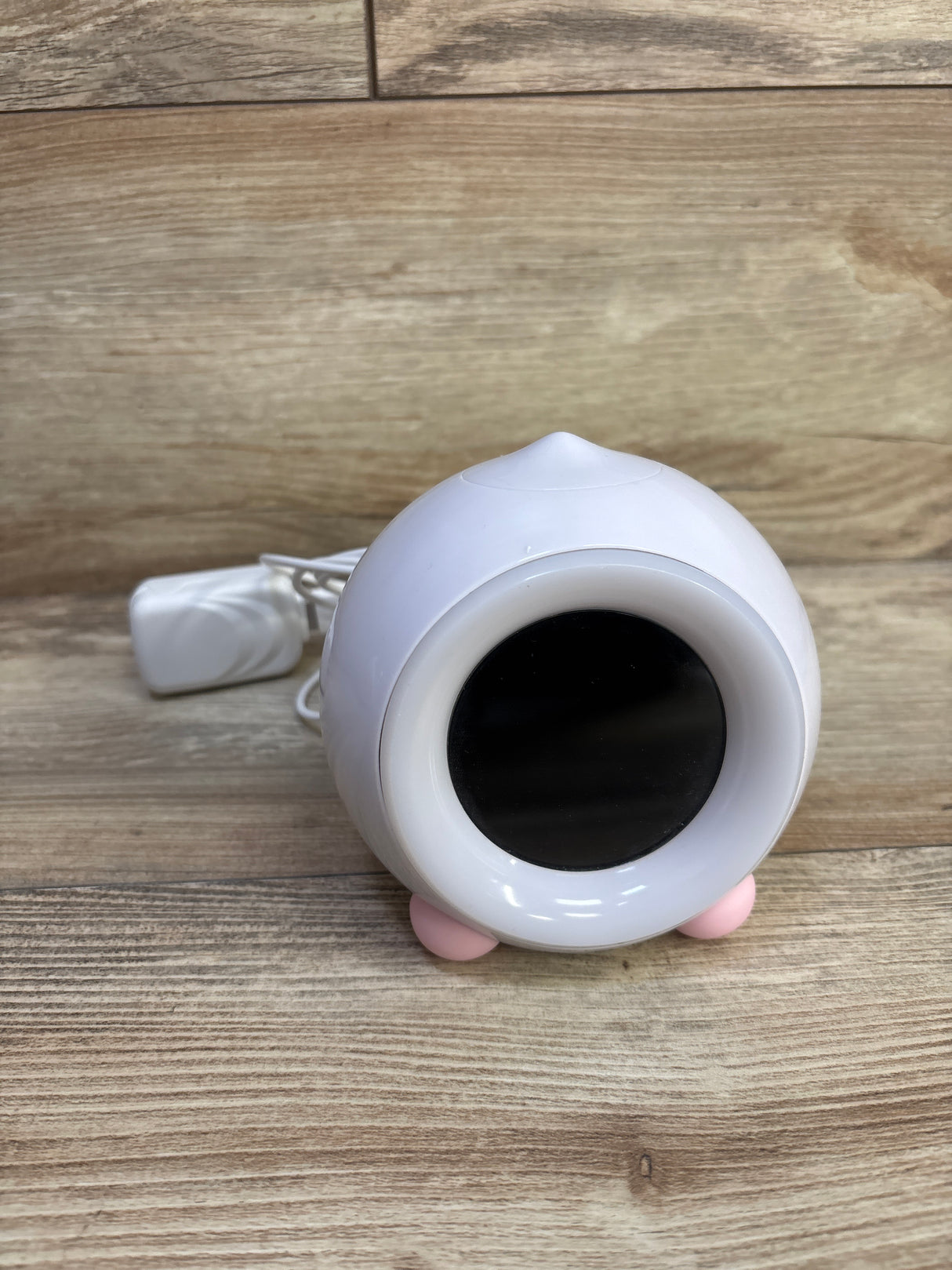 Angled front of LittleHippo Mella Ready To Rise sleep trainer showing black display, pink feet, and power cord on wood