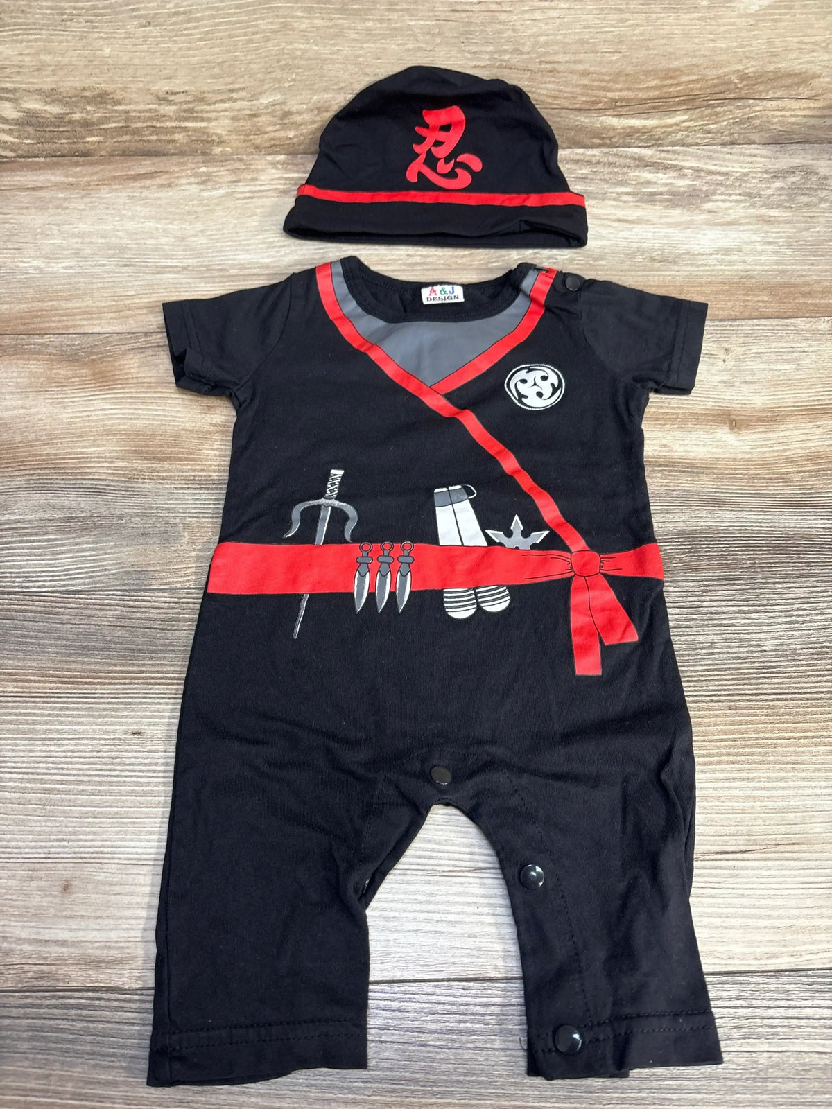 Flat lay of black baby ninja romper and hat showcasing red sash, ninja graphics, and snap leg closures on wood
