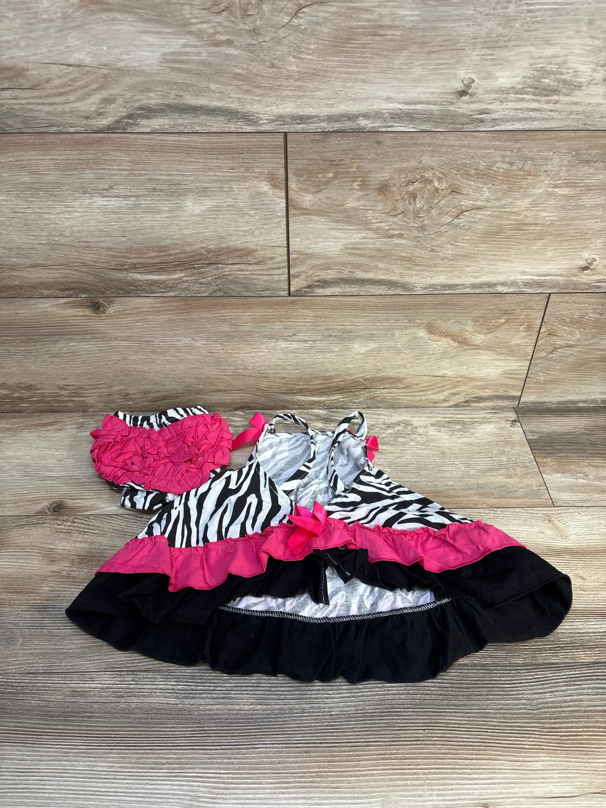 Flat lay of zebra-print open-back baby top and matching ruffled bloomers showing hot pink and black ruffle trim on wood floor