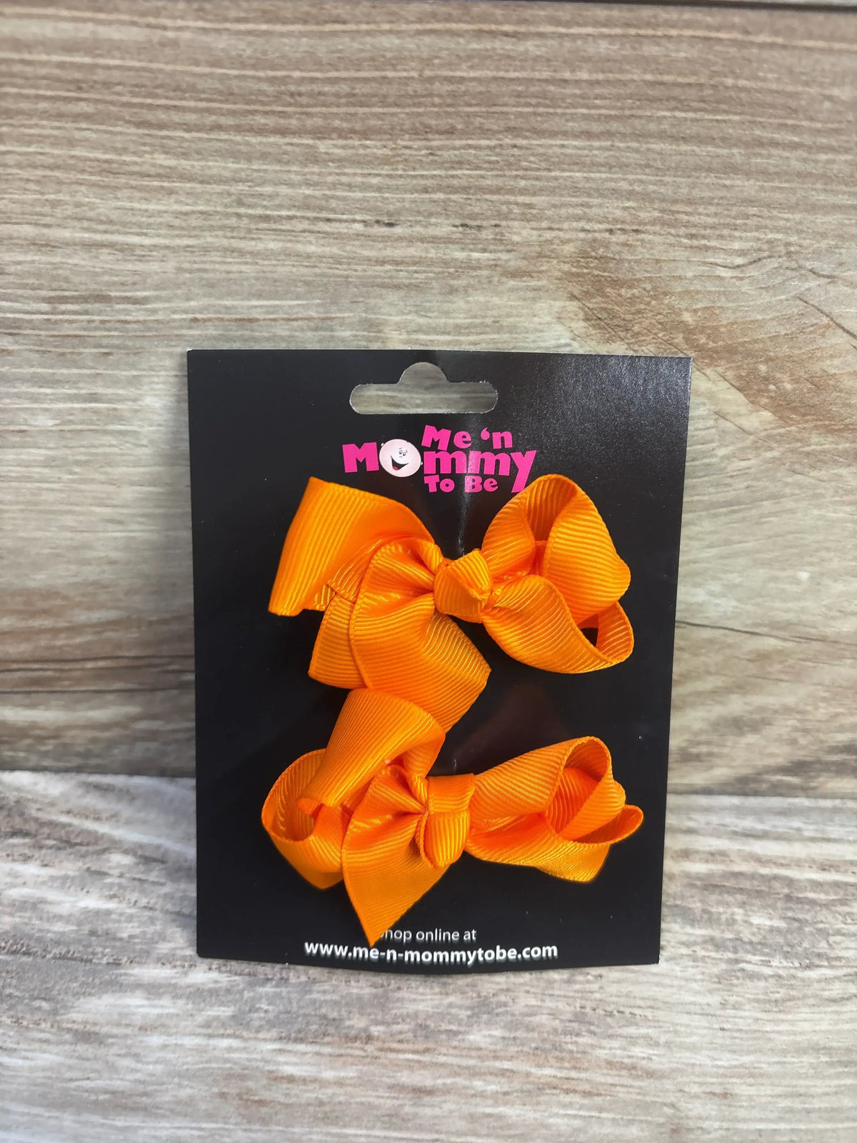 Front view of two orange grosgrain hair bows with clips on a black branded card over light wood background