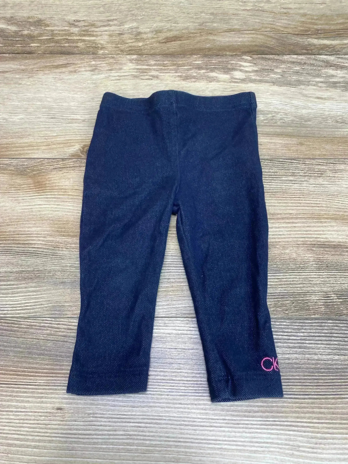 Flat lay of Calvin Klein leggings size 12 months showing blue denim-look knit and pink CK logo at left hem on wood floor