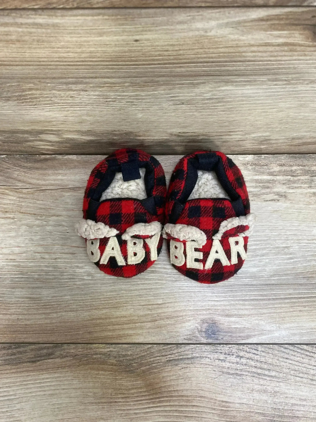 Top view of baby buffalo-plaid bear slippers showcasing cream BABY BEAR letters and sherpa lining on light wood floor