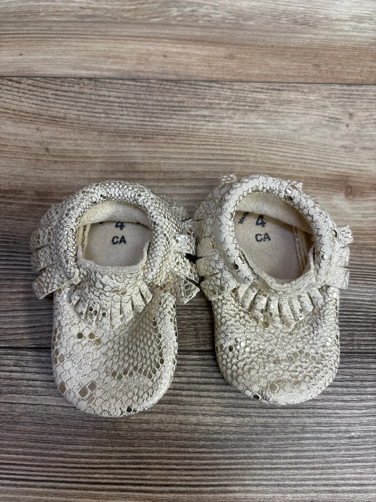 Top view of Freshly Picked snakeskin moccasin sneakers size 4C showcasing metallic beige snakeskin texture and fringe on wood floor