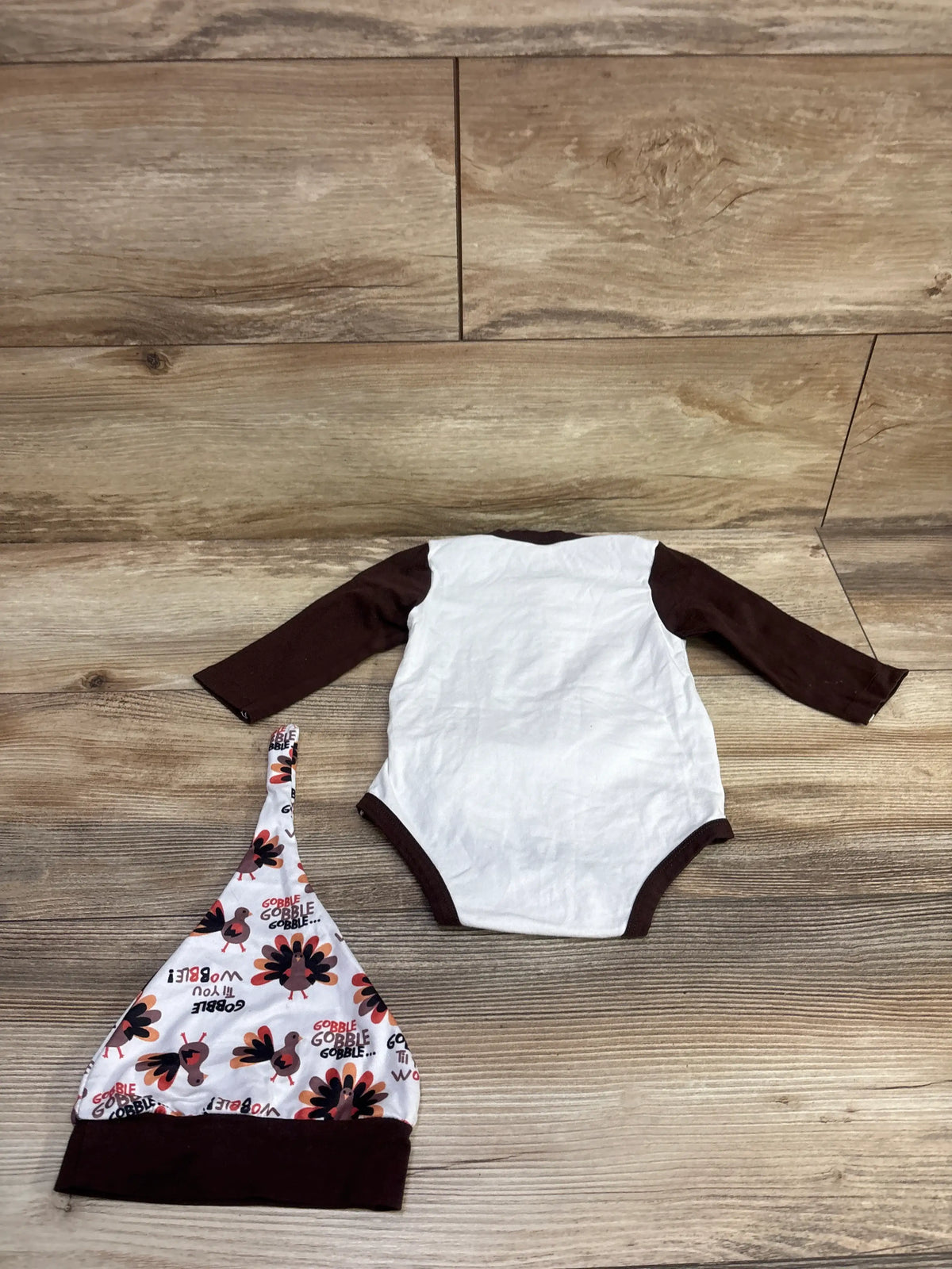 Top view of Thanksgiving faux suspender bodysuit set showcasing brown trim and matching turkey-print hat on wood flooring
