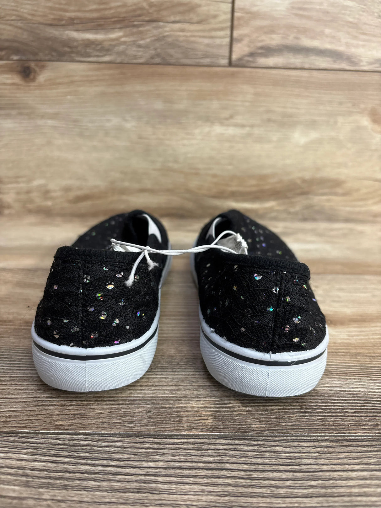 Pair of black kids' slip-on shoes with white soles and iridescent dots on wood floor background
