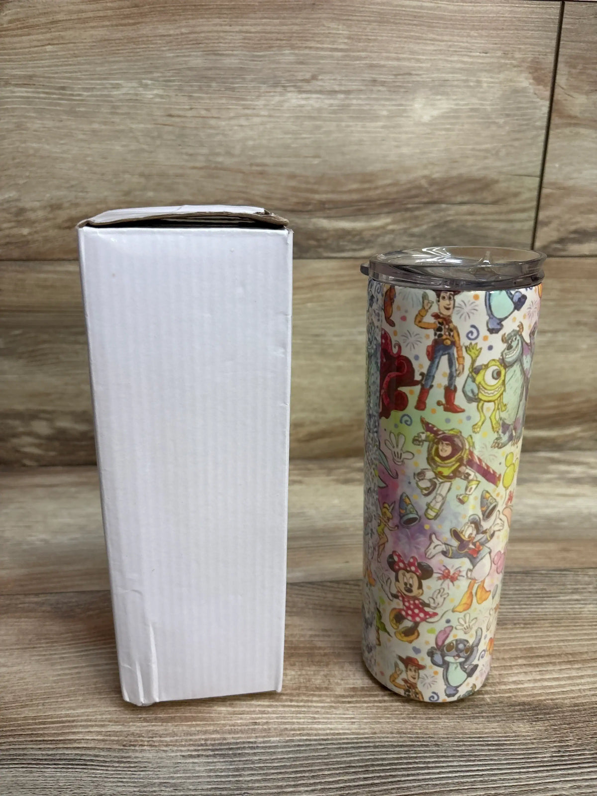 Tall tumbler with colorful Disney characters including Woody, Buzz Lightyear, Mickey and Minnie Mouse next to a plain white box on wooden surface