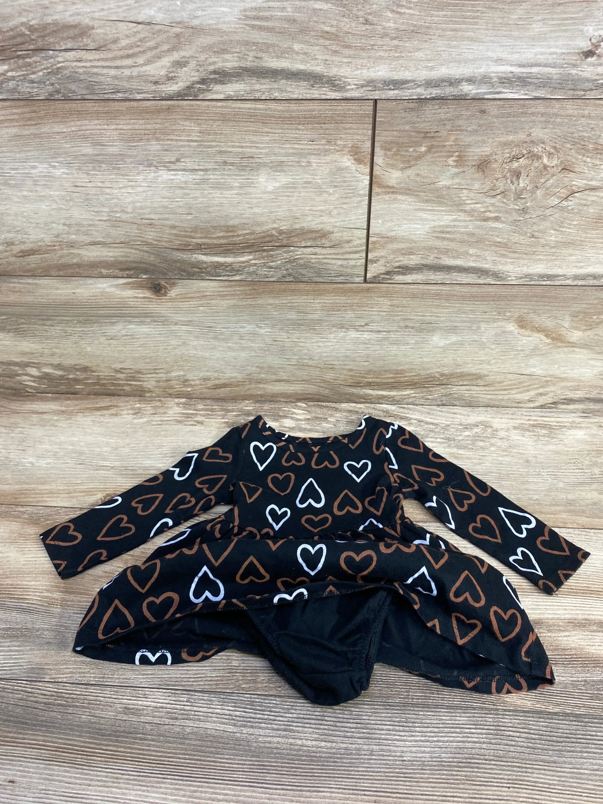 Black long-sleeve baby dress with brown and white heart patterns, laid flat on wooden floor