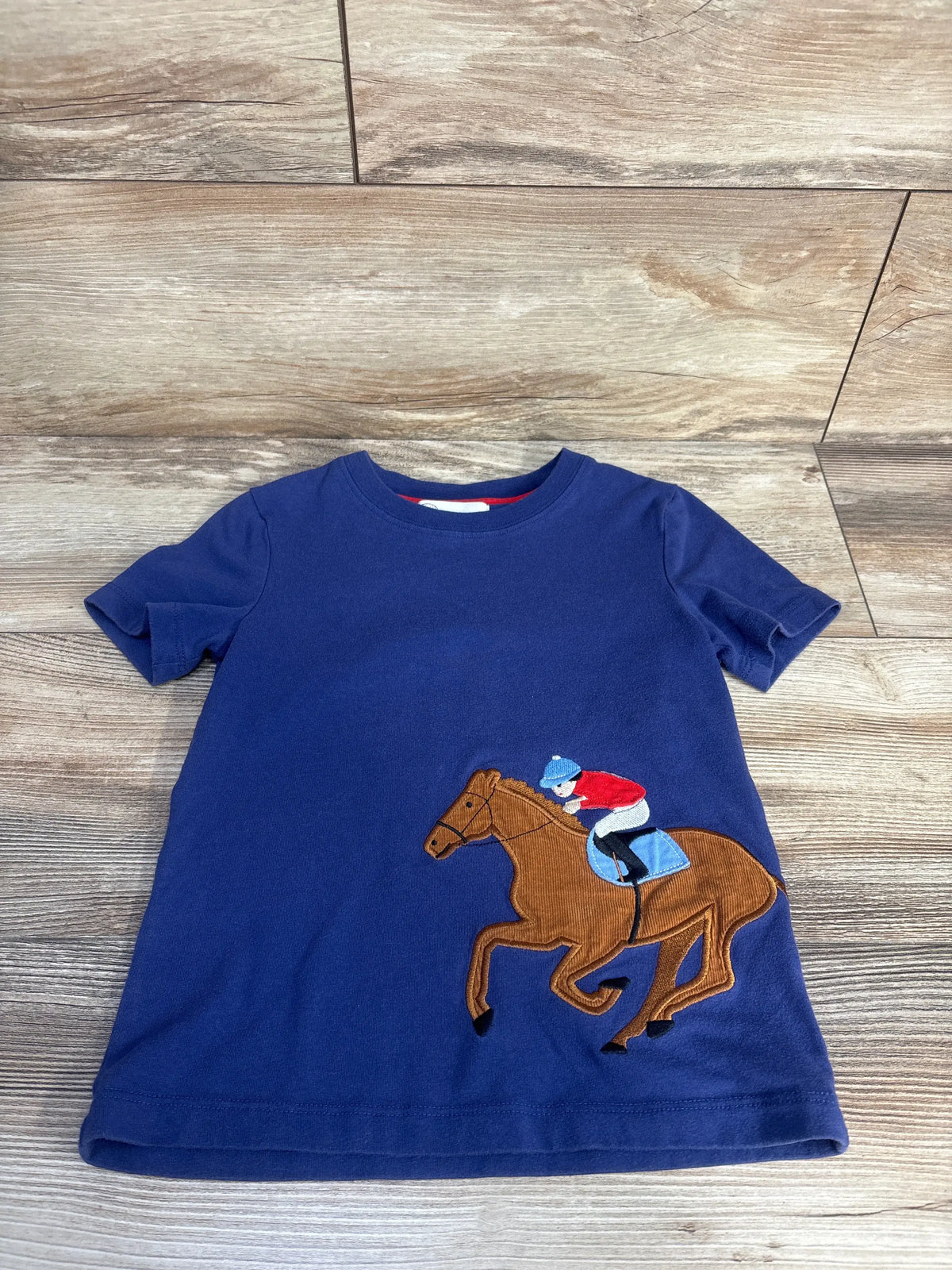 Front flat lay of blue 5T short-sleeve shirt with embroidered brown horse and jockey on light wood background