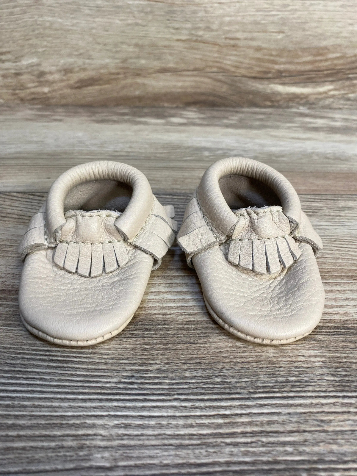 Cream leather baby moccasins with fringe detail on wood background, kids shoes resale