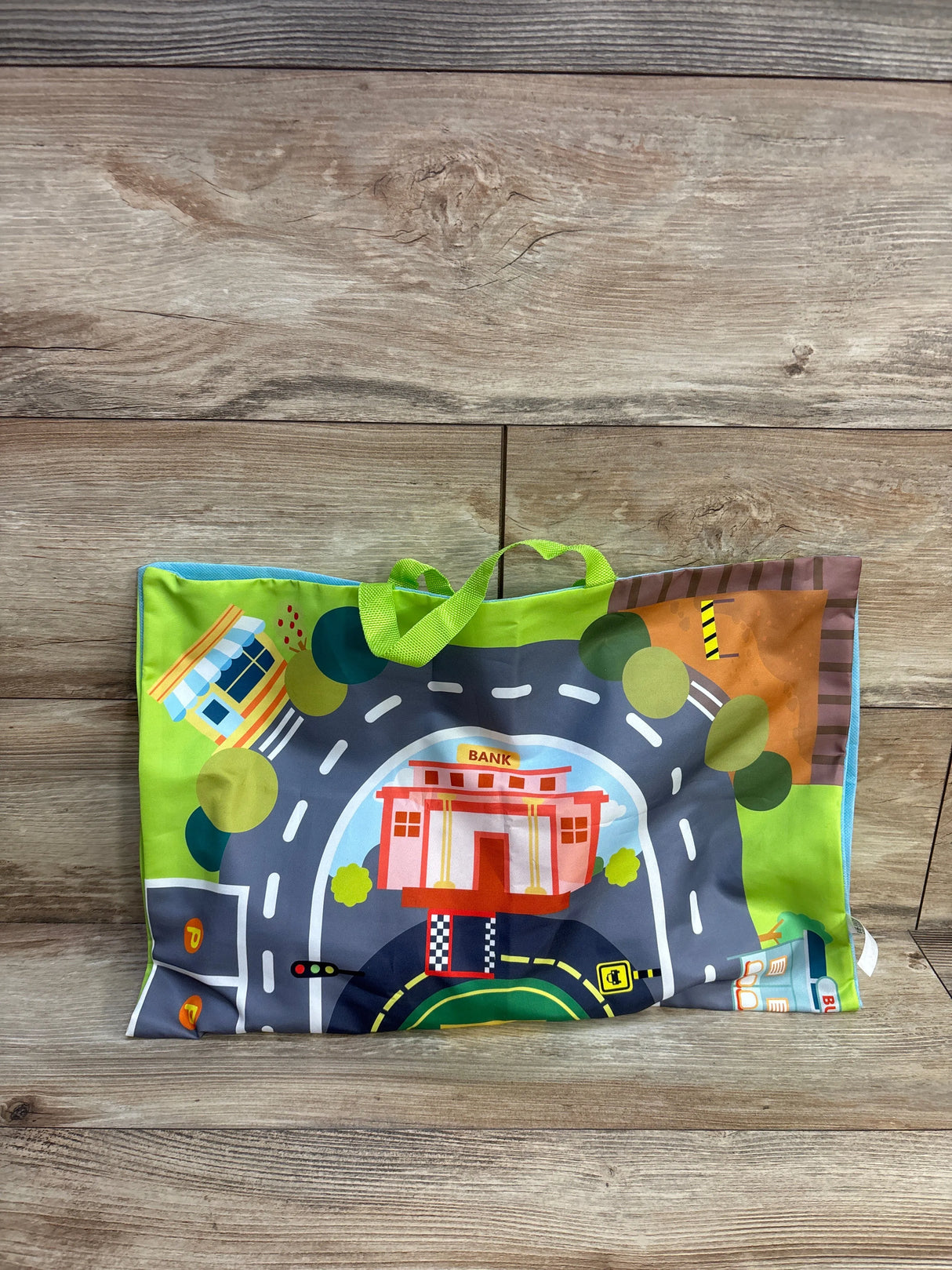 Front view of folded playmat showing printed road, bank and buildings with green carrying handles on wood background