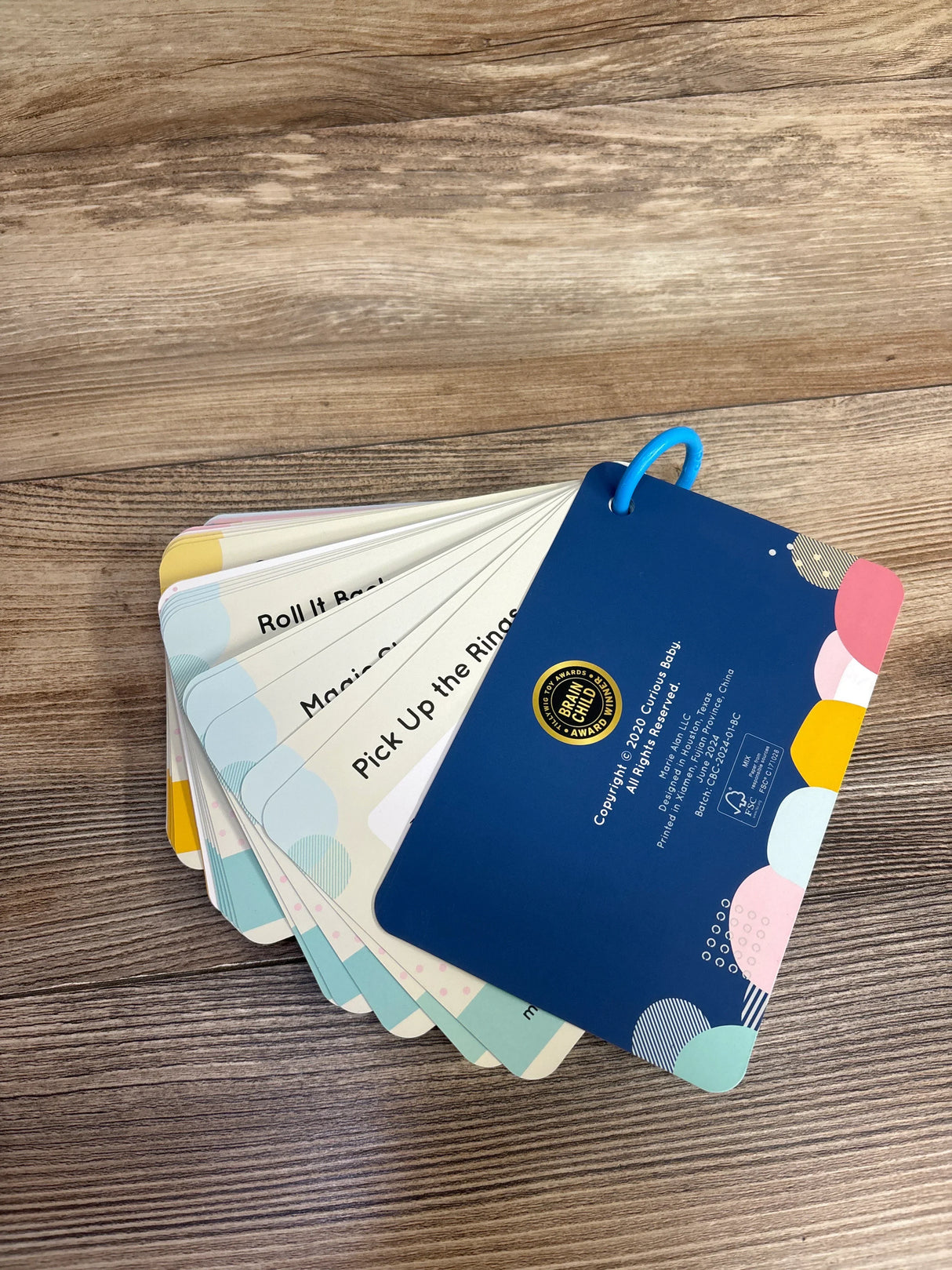 Angled top-down view of Curious Baby Activity Cards fanned on a wooden table, blue ring binding and visible prompt Pick Up the Rings