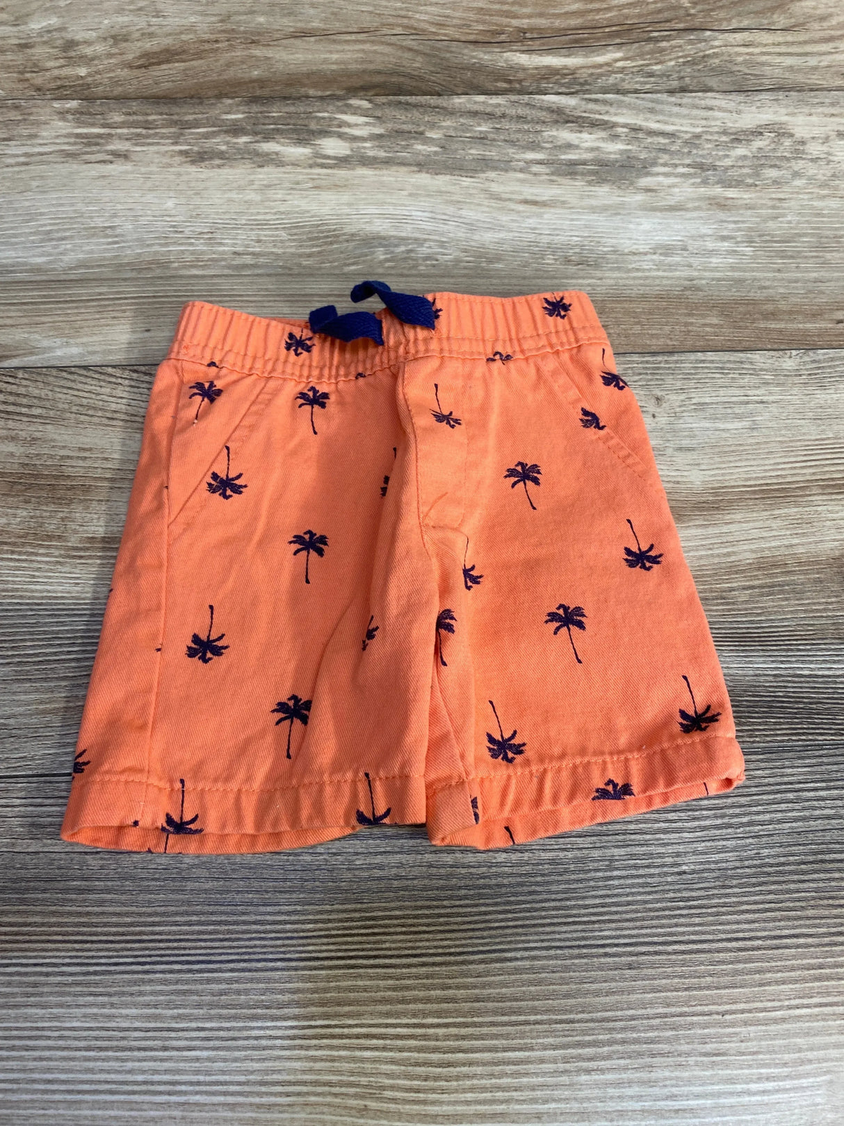 Top view of orange baby shorts 6-9m showing navy palm-tree print and elastic waist with navy drawstring on wood surface