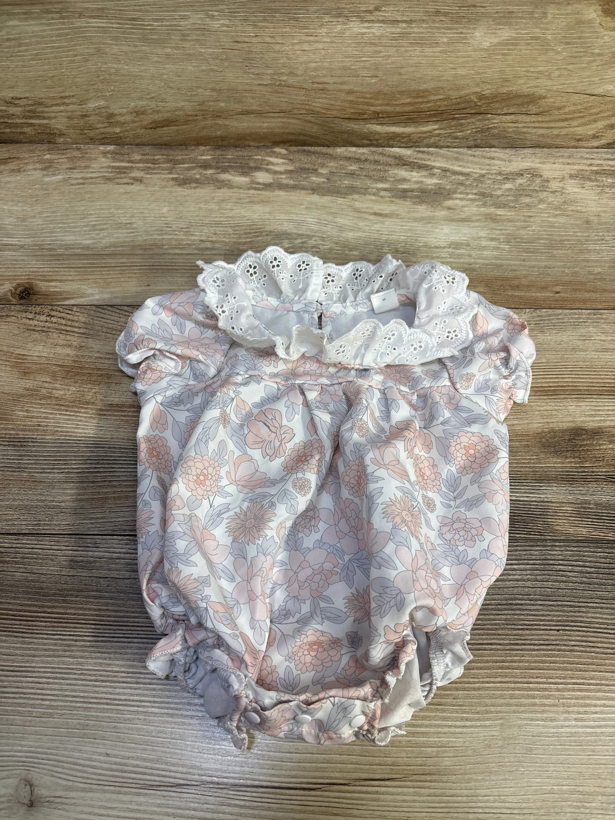 Top-down view of white baby bubble romper with eyelet ruffle collar and pink floral print on wood background