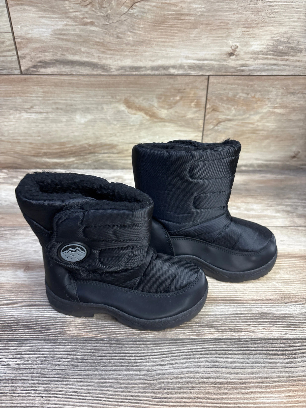Three-quarter side view of a pair of black toddler snow boots with quilted nylon shaft, faux-fur lining, and rubber soles on wood background