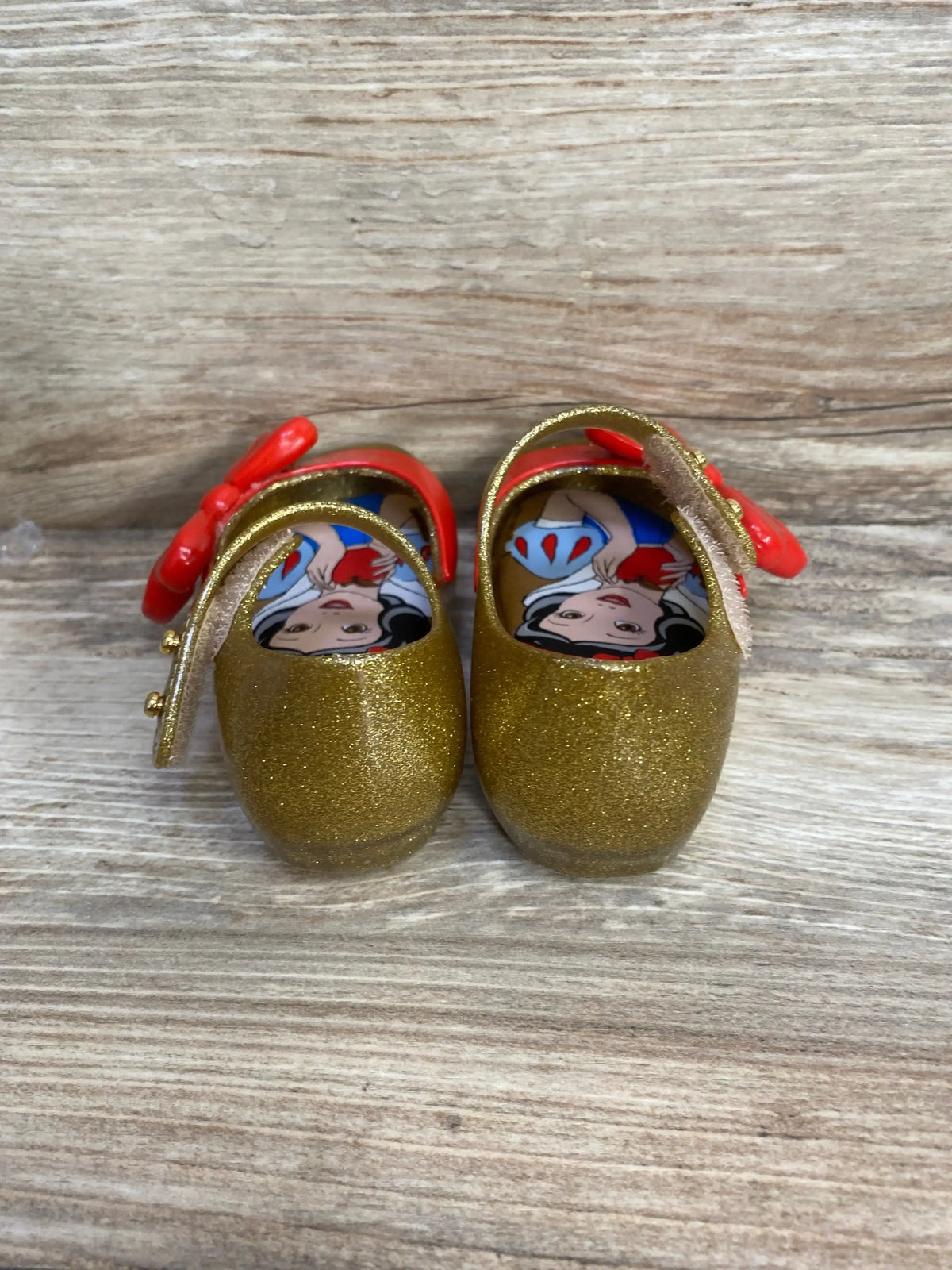 Front view of toddler gold glitter Ultragirl Mary Jane shoes showing Snow White insole and red bows on wood surface