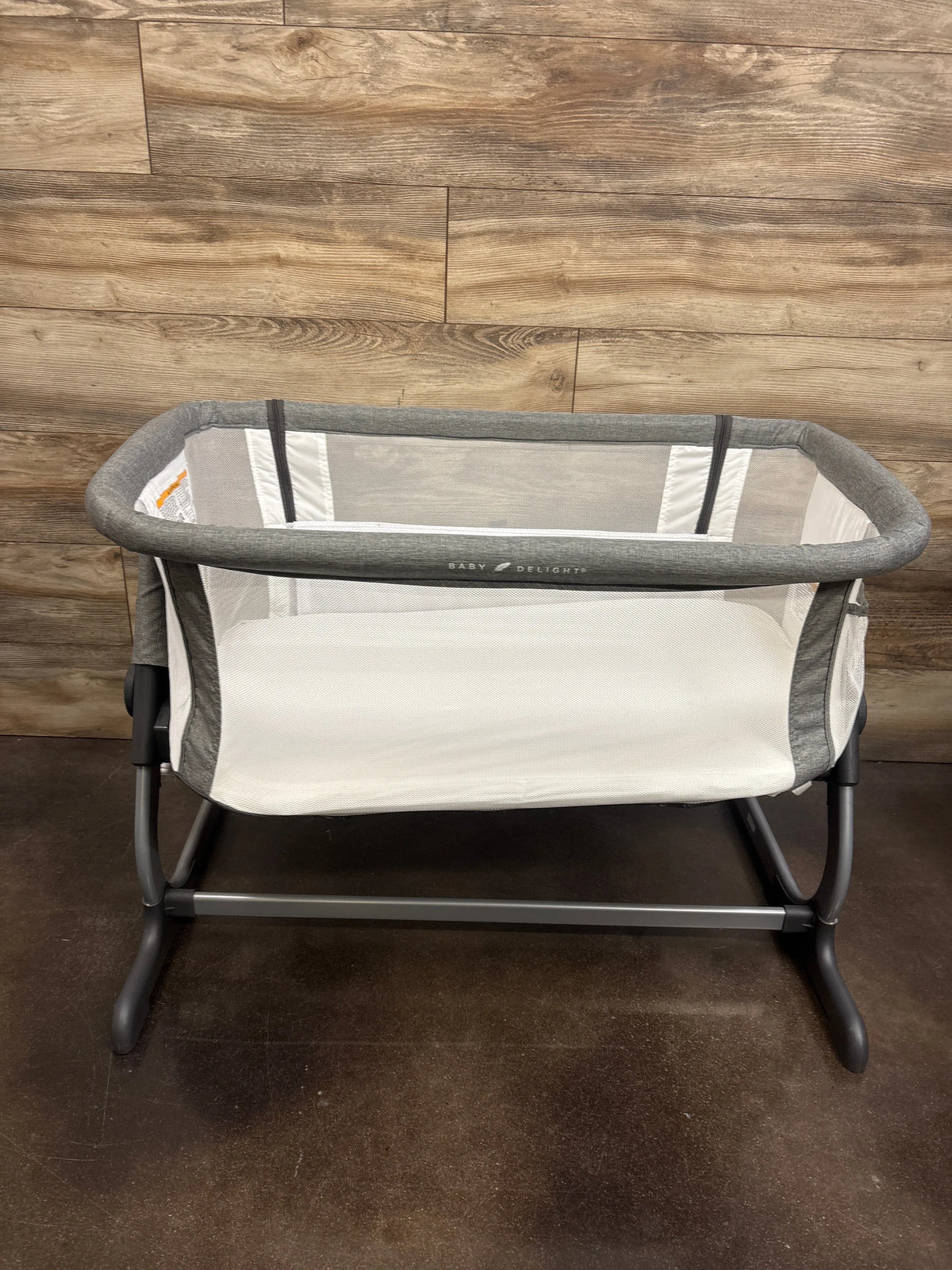 Front view of Baby Delight Beside Me Dreamer bedside bassinet in grey showcasing mesh sides with a wood floor background