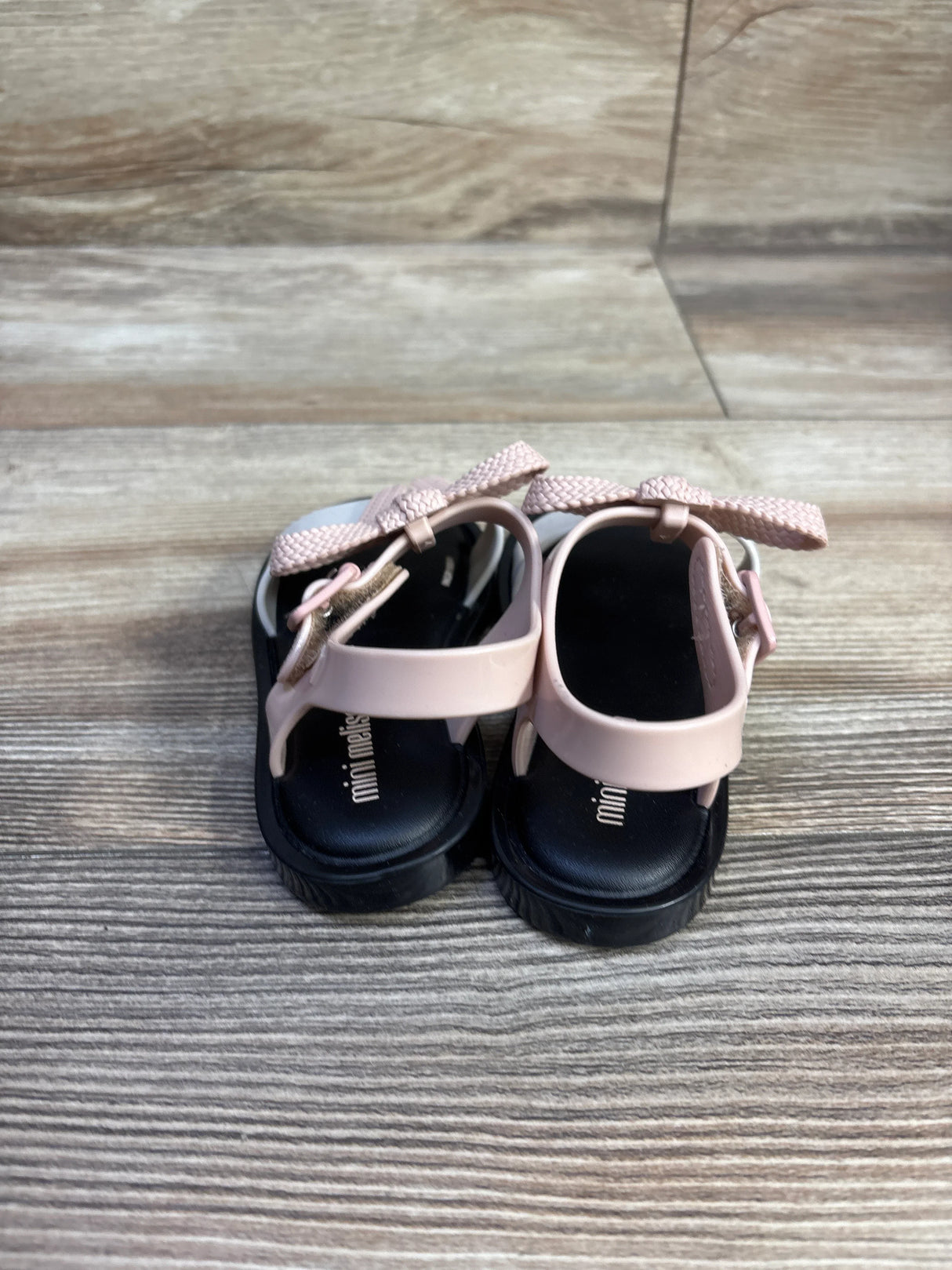 Back view of Mini Melissa Mar toddler sandals showcasing pale pink braided bow straps and black footbeds on wood background