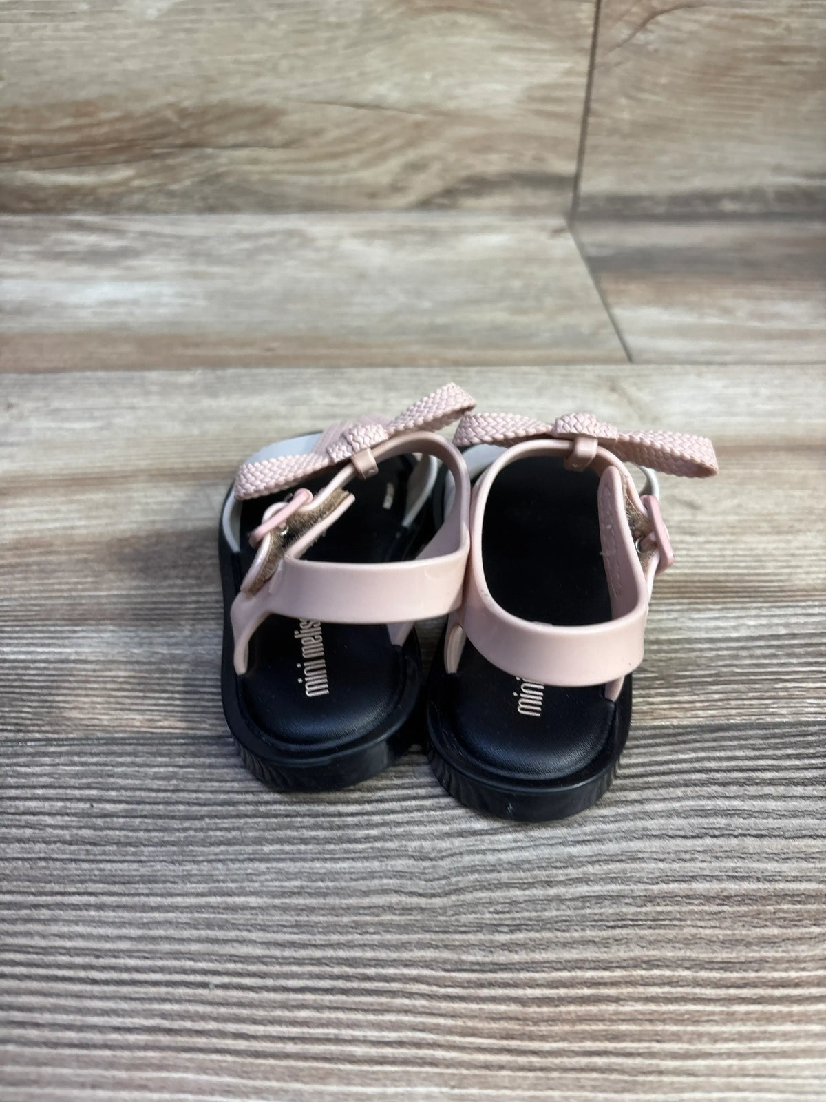 Back view of Mini Melissa Mar toddler sandals showcasing pale pink braided bow straps and black footbeds on wood background