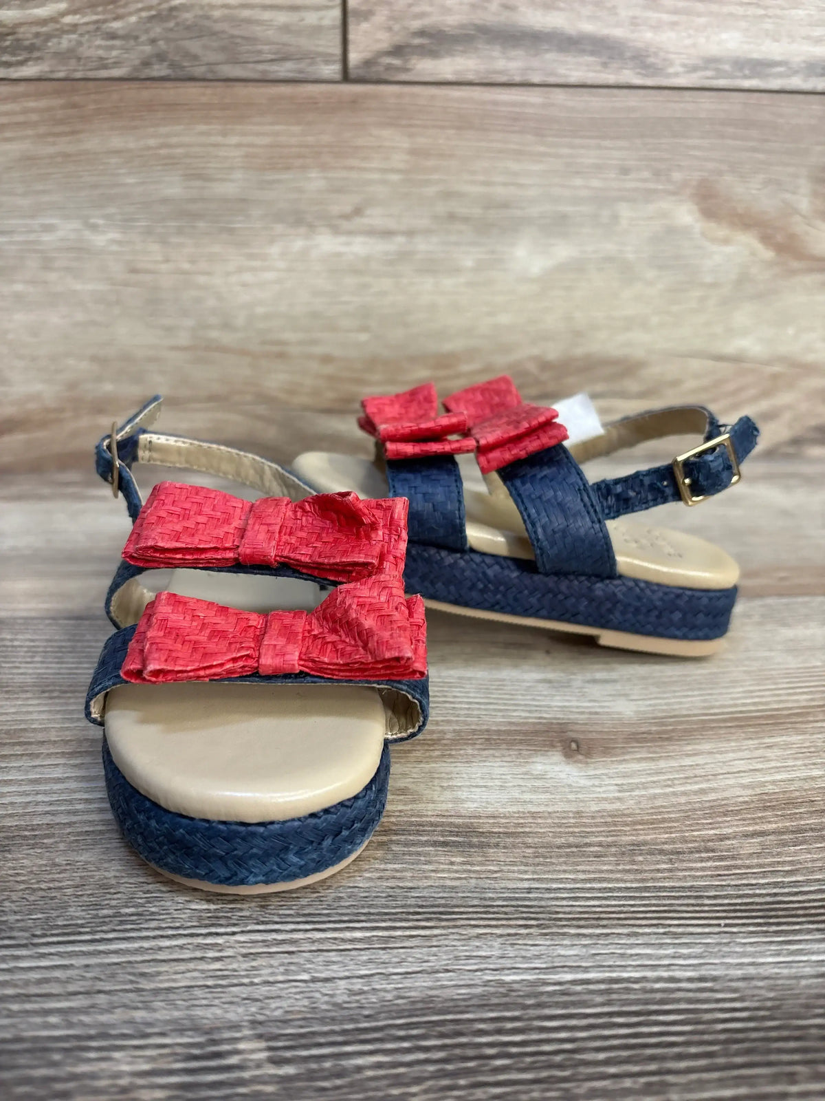 Angled view of Janie and Jack double bow straw sandals in Merchant Marine sz 6c showcasing navy woven straw platform with red raffia bows on wood plank floor