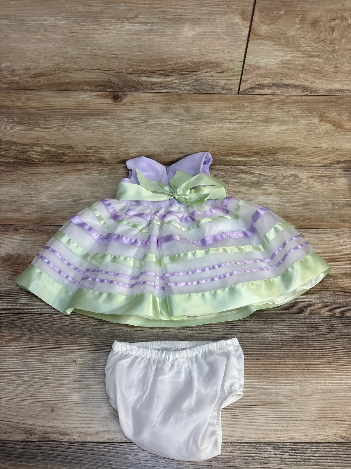 Pastel purple and green baby dress with ruffles and bow, paired with white bloomers on wood floor