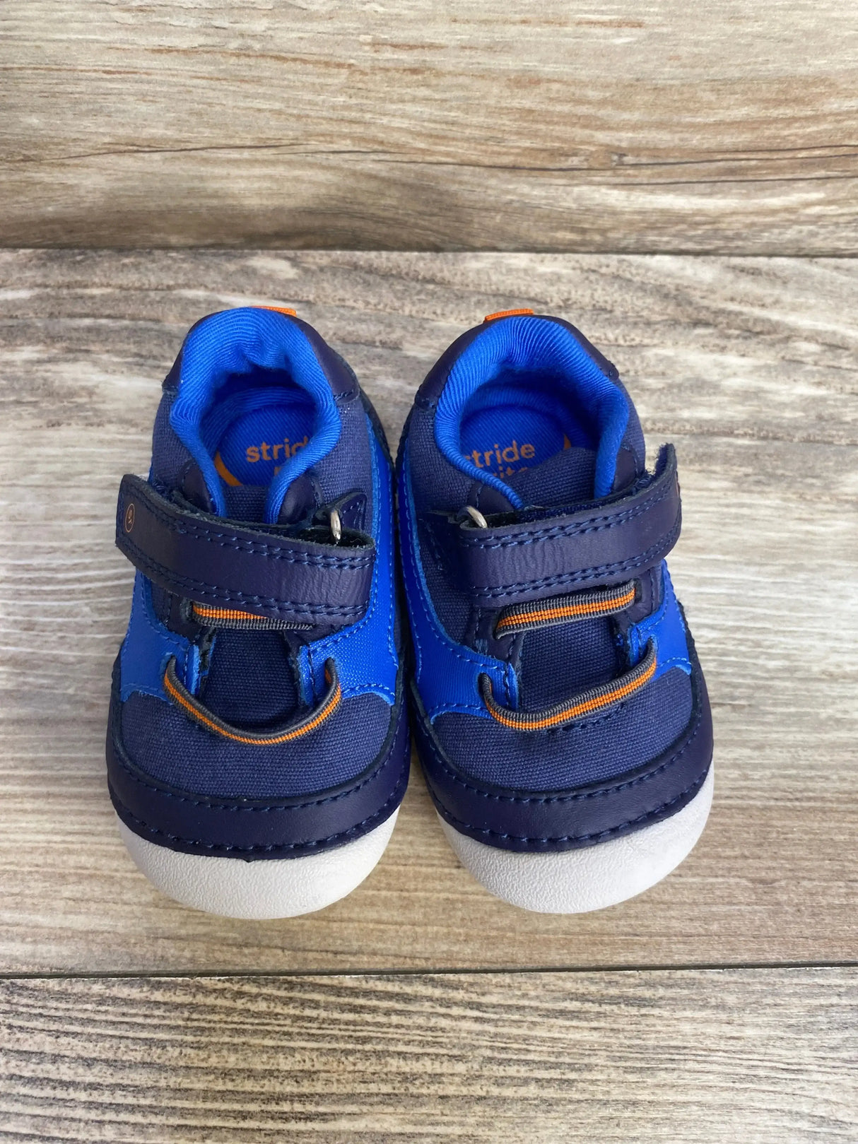 Top-down view of Stride Rite Soft Motion Kylin toddler sneakers showing navy and blue uppers, orange trim, velcro straps, and white rounded toe caps on wood background