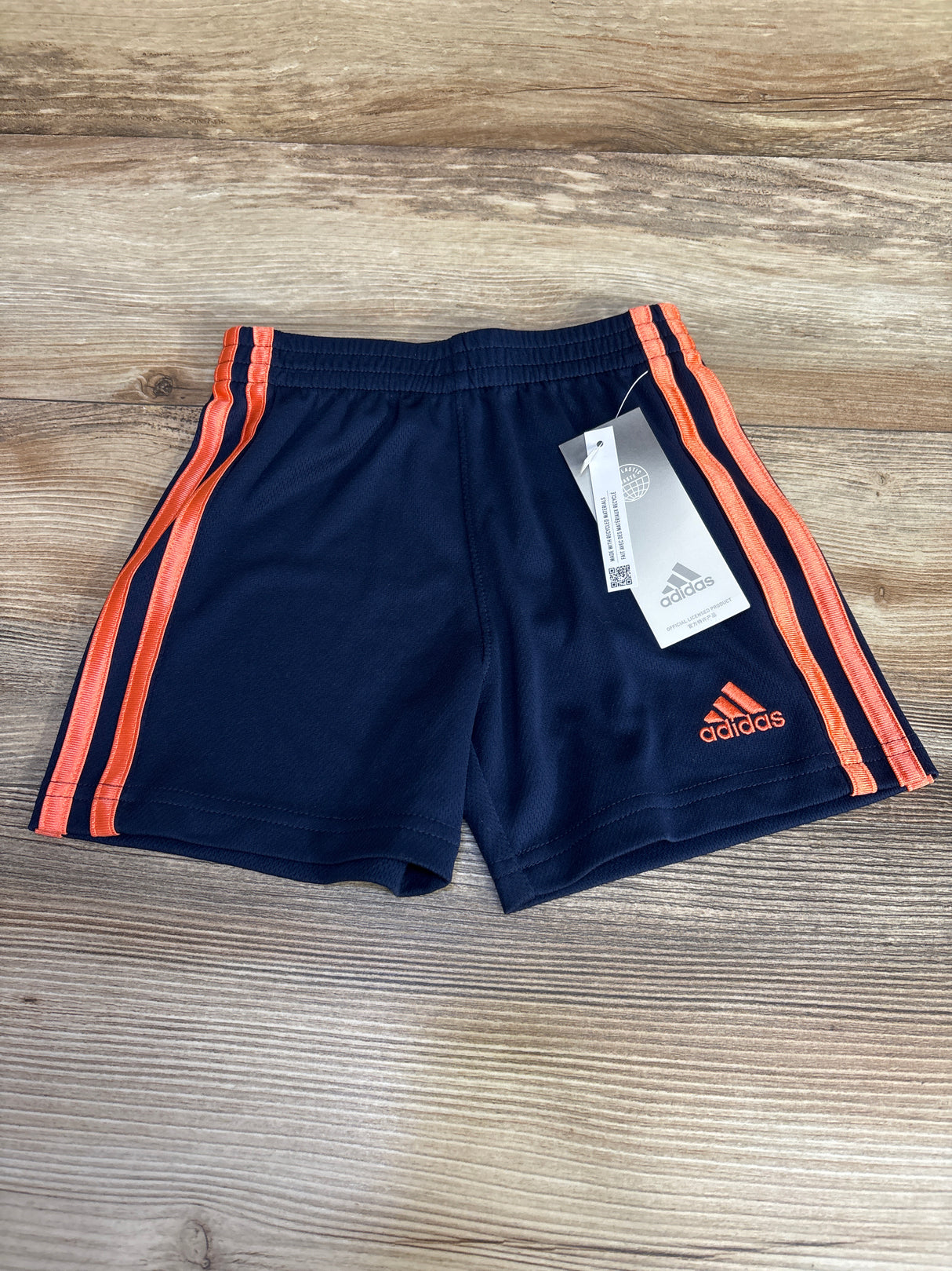 Navy blue Adidas kids shorts with orange stripes and logo, displayed on wood background