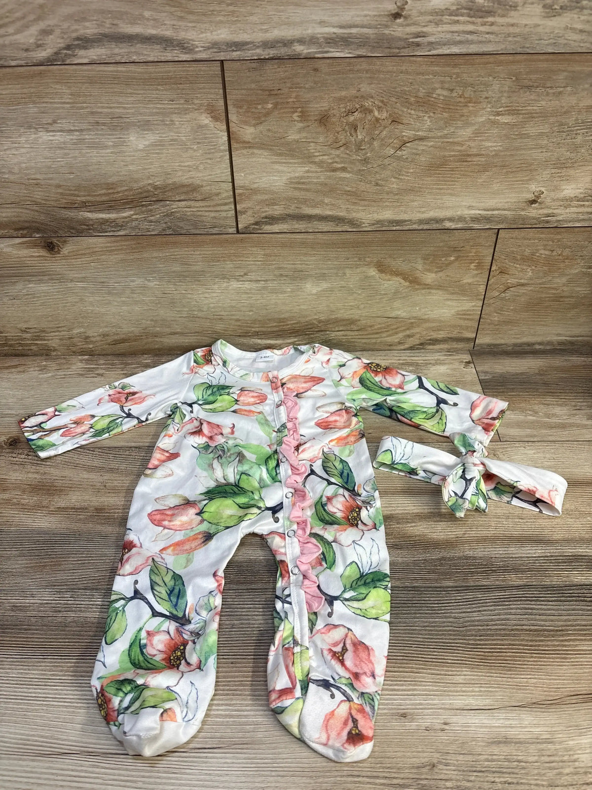 Flat lay of white floral footed sleeper and matching headband showcasing ruffle trim and front snap closures on wood floor