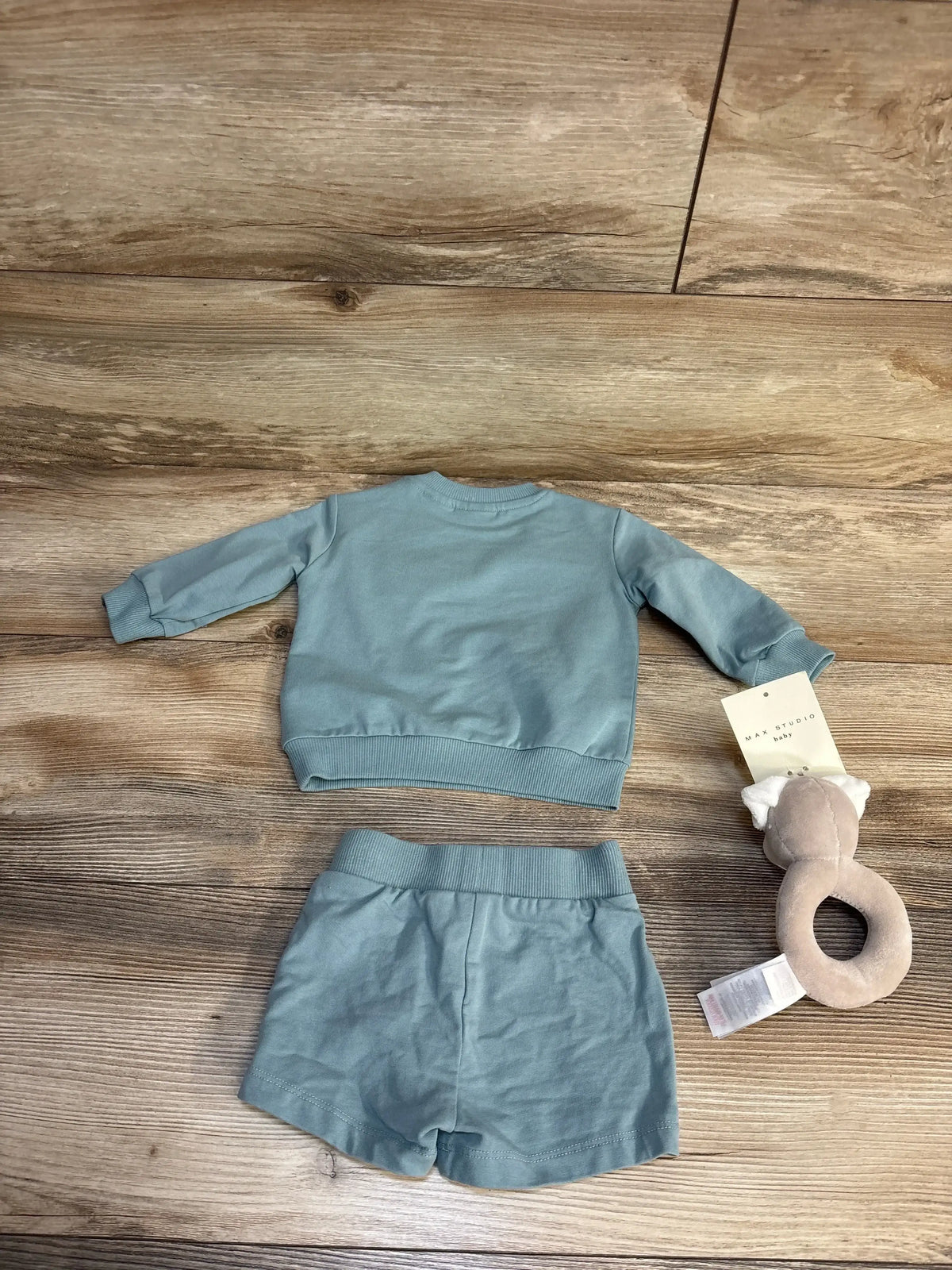 Top-down view of Max Studio baby 3pc sweatshirt, shorts and rattle set in blue on a wood floor