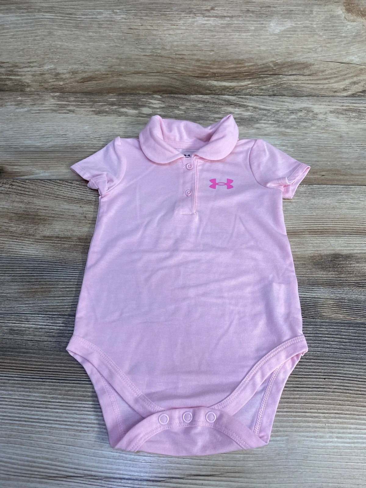 Flat lay of pink Under Armour baby polo bodysuit showing Peter Pan collar, two-button front placket, pink logo and three snap crotch on wood background