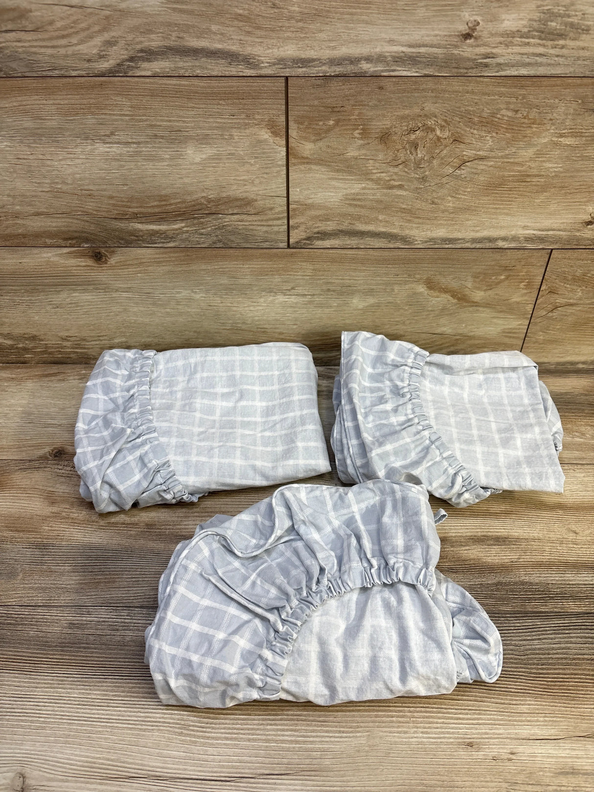 Top view of three light gray and white windowpane fitted bassinet sheets showing elasticized corners on a wood plank background
