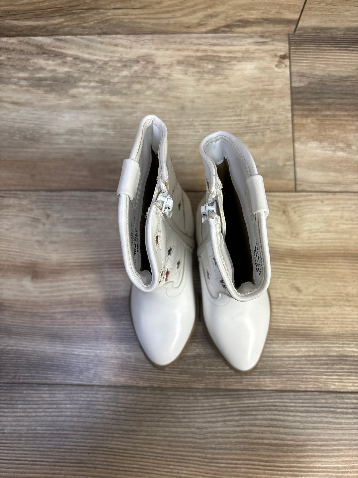 Top-down view of white toddler cowgirl boots showing pointed toes, side zippers, star cutouts with pink lining on wood floor
