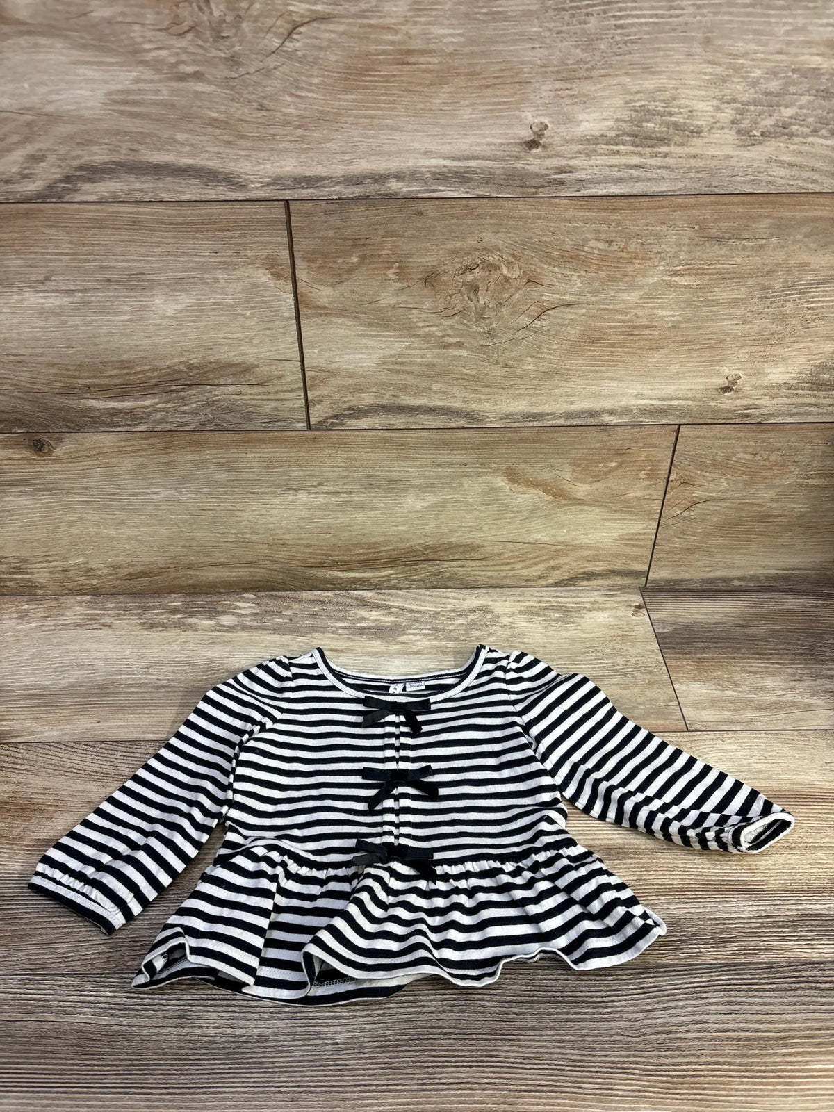 Flat-lay front of Janie & Jack striped peplum top showing three black bows and long sleeves on wood floor background
