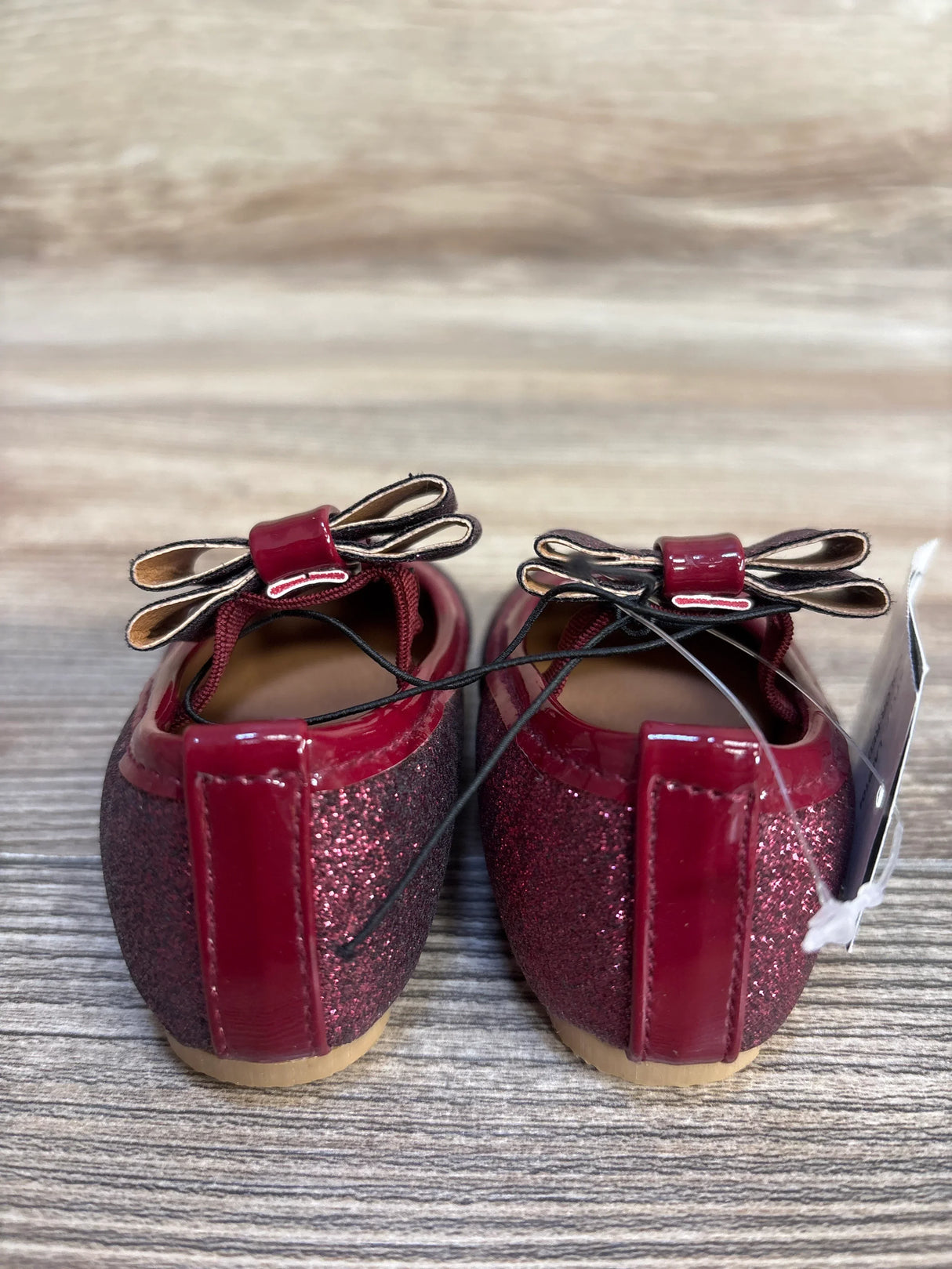 Shiny burgundy baby shoes with glitter, bow details, and elastic laces on wood floor