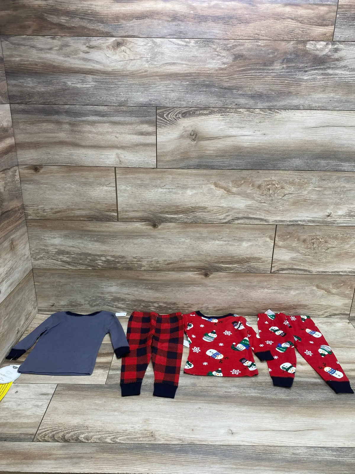 Flat-lay of Koala Kids 4-piece pajama set showing gray long-sleeve top, red buffalo-plaid pants, and red snowman set on wood floor