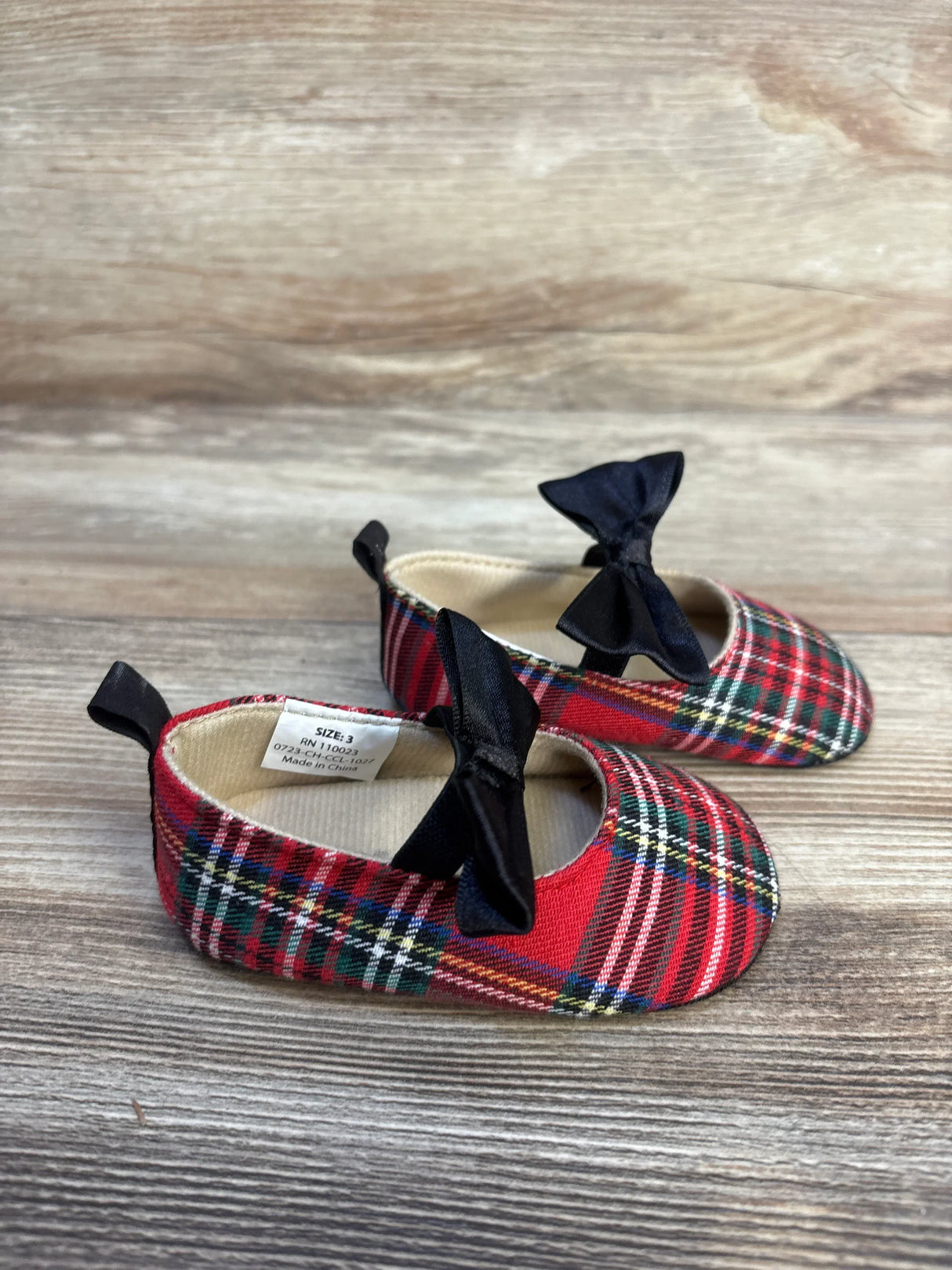 Red plaid baby shoes with black bows on wood background, toddler footwear