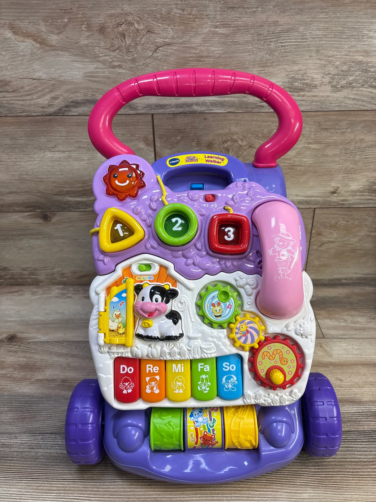 Front of VTech Sit-to-Stand walker with purple handle, activity panel with shape buttons and piano keys on wood floor