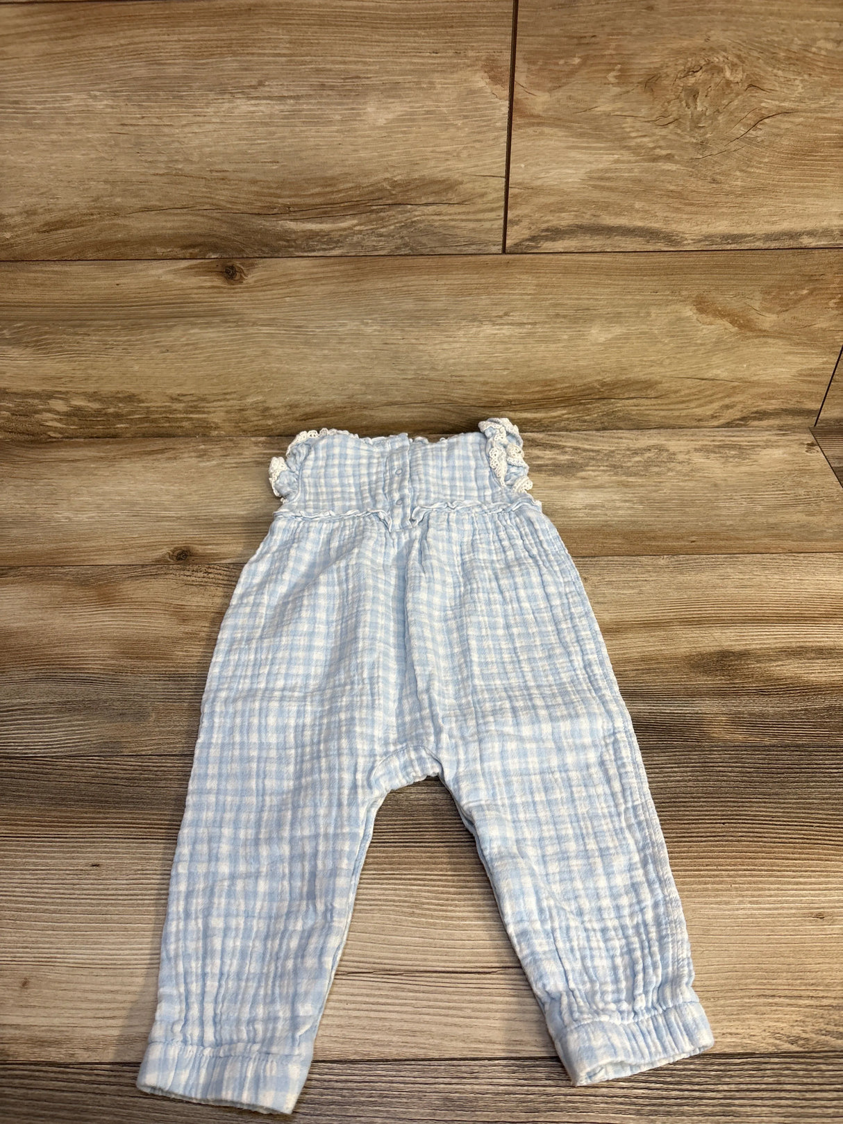 Flat lay of baby gauze jumpsuit in light blue check showing smocked ruffled top and elastic cuffs on wood background