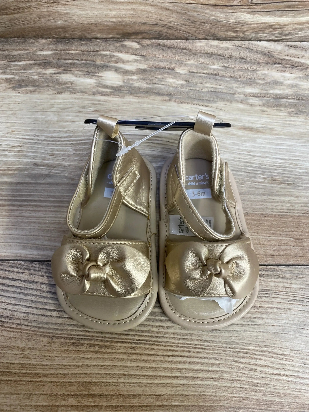 Top view of Child of Mine gold baby sandals showcasing front bow detail and Velcro strap on wooden background