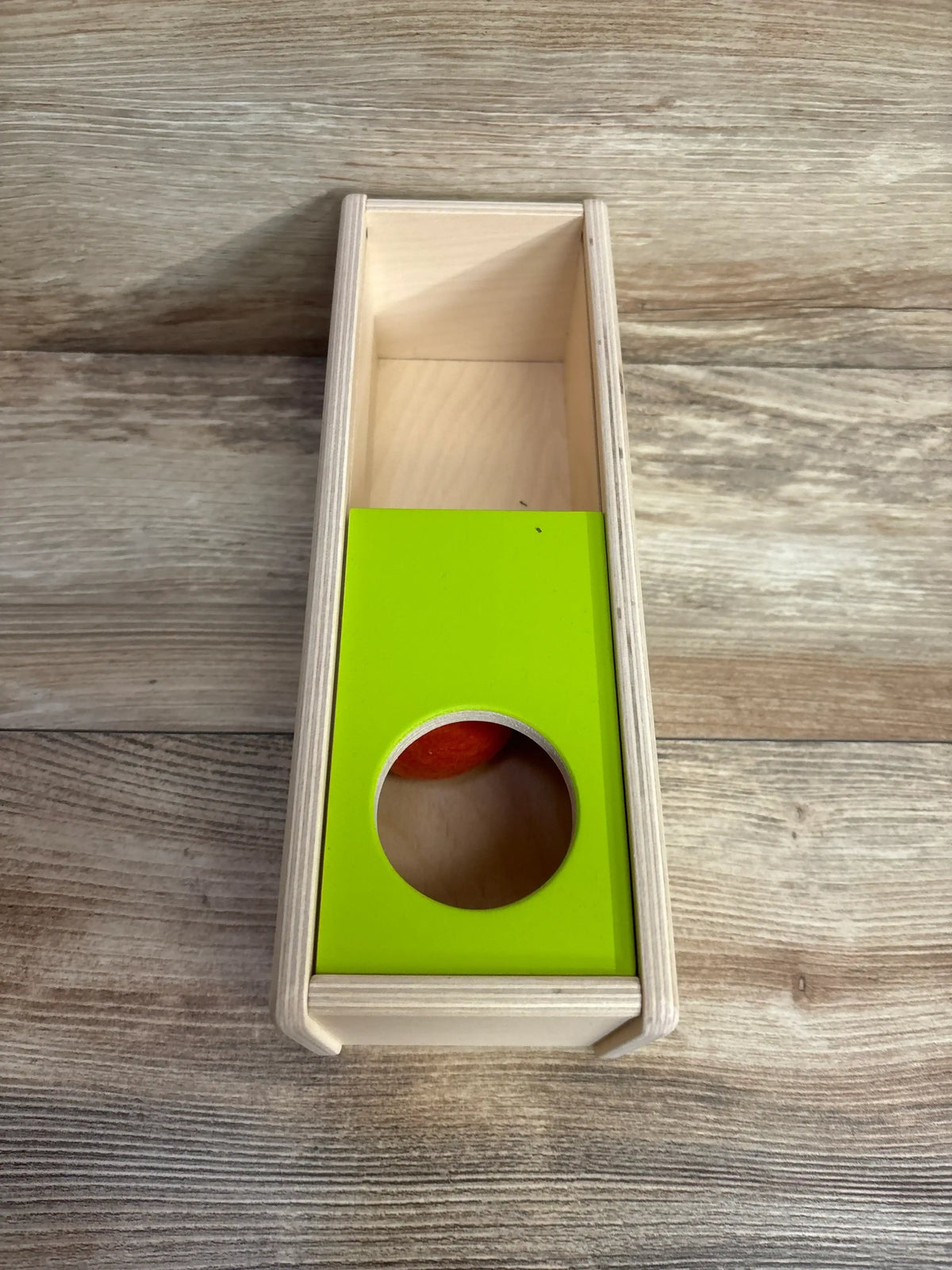 Top-down view of Lovevery Sliding Top Box showing green sliding top with circular hole and orange felt ball inside on wood floor