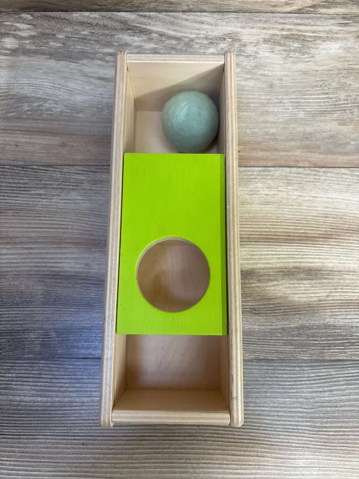 Top view of Lovevery Sliding Top Box with bright green sliding panel, circular opening, and gray ball on wood-grain surface