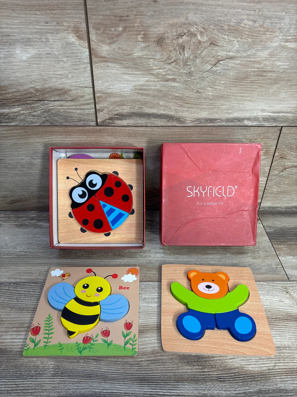Top-down view of Skyfield 4 pack wooden animal puzzles showing colorful ladybug, bee, and bear pieces next to a red box on wood surface