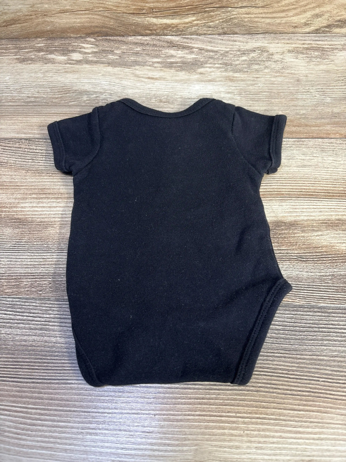 Back view of black newborn short-sleeve bodysuit showing plain cotton fabric and leg openings on wood plank background