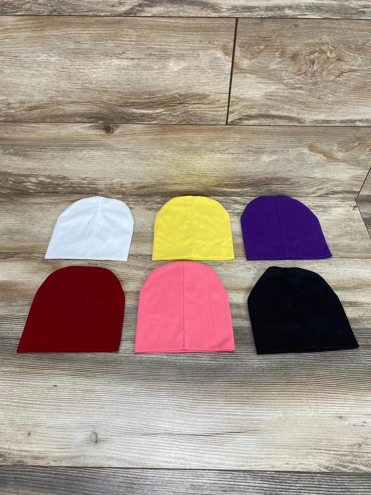 Top-down view of 6-pack basic baby beanies showcasing assorted bright colors laid flat on wood background