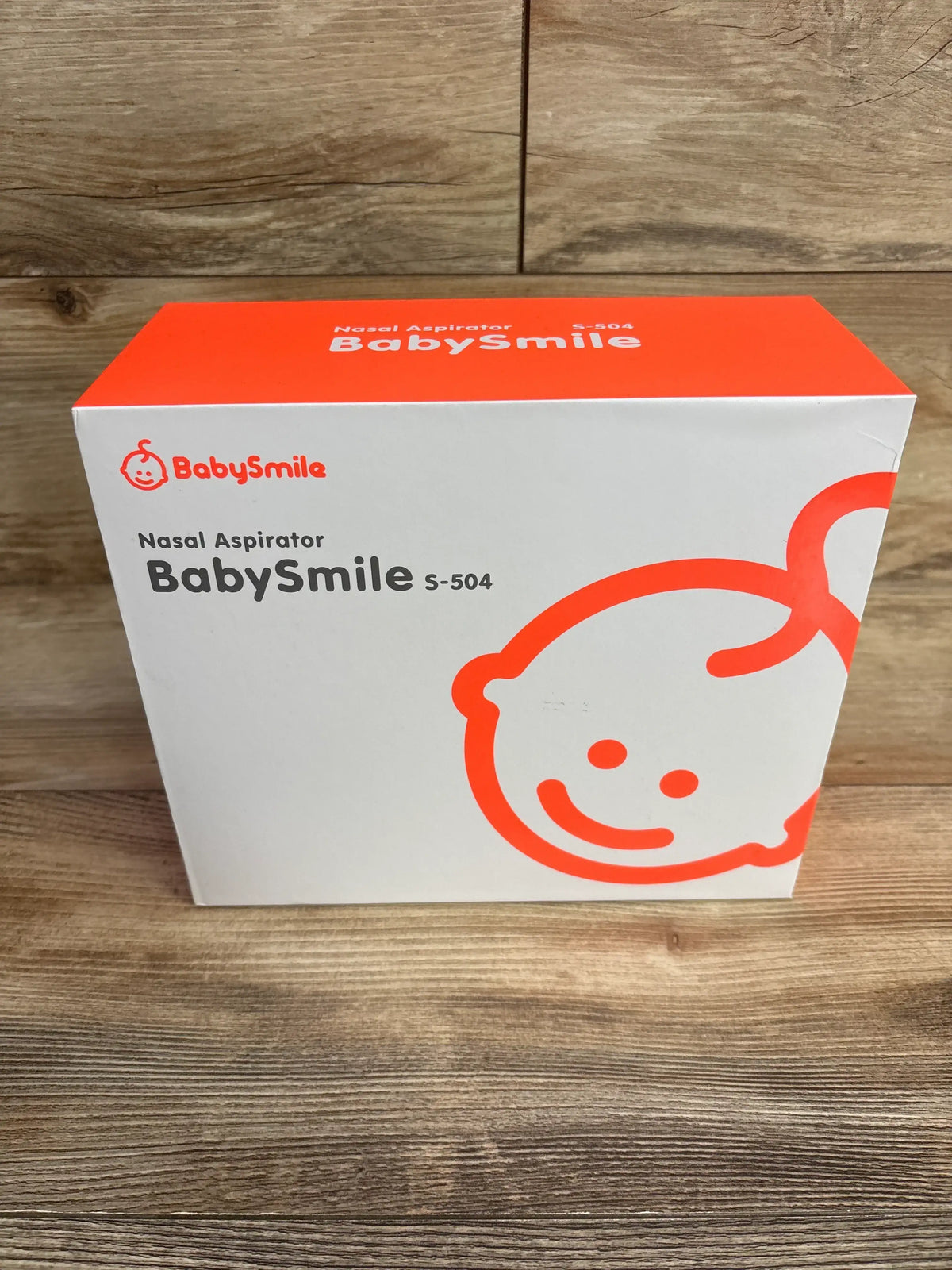 Front view of BabySmile S-504 nasal aspirator box showcasing orange baby face graphic and brand name with wooden background