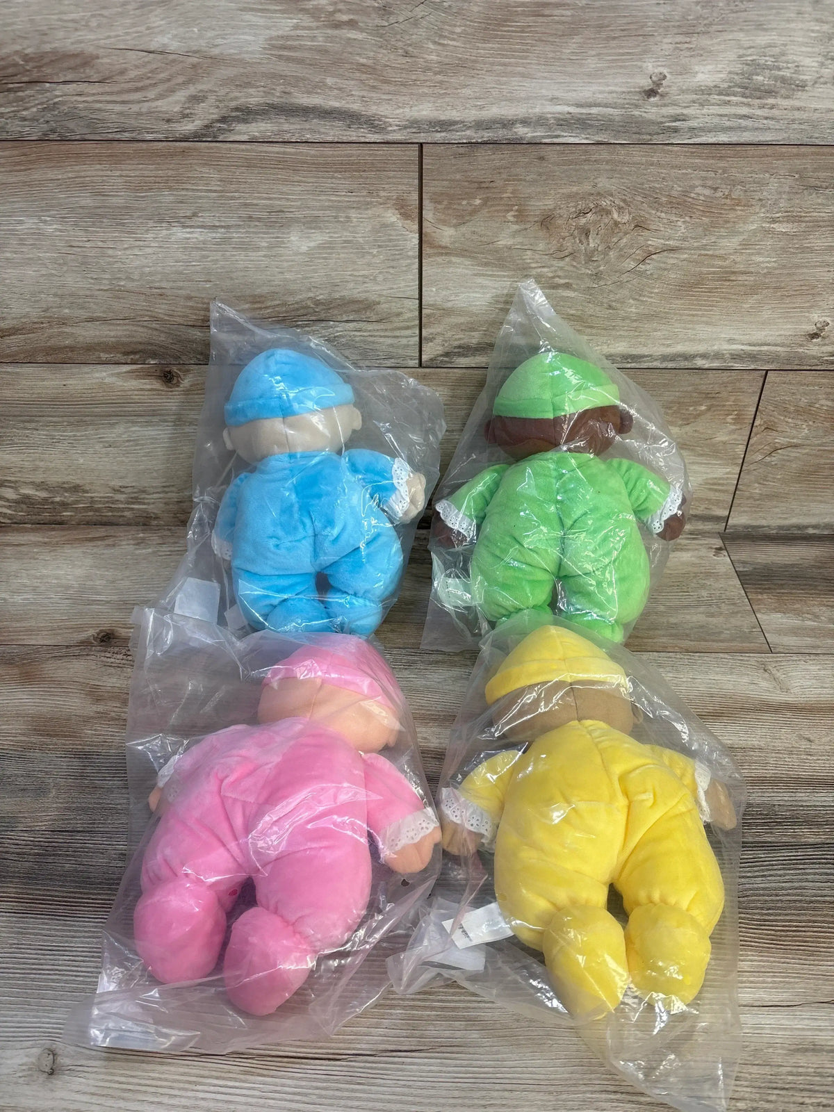 Flat lay of NEW 4pc Lakeshore Cuddly Washable Dolls in blue green pink and yellow outfits sealed in plastic bags on wood plank floor