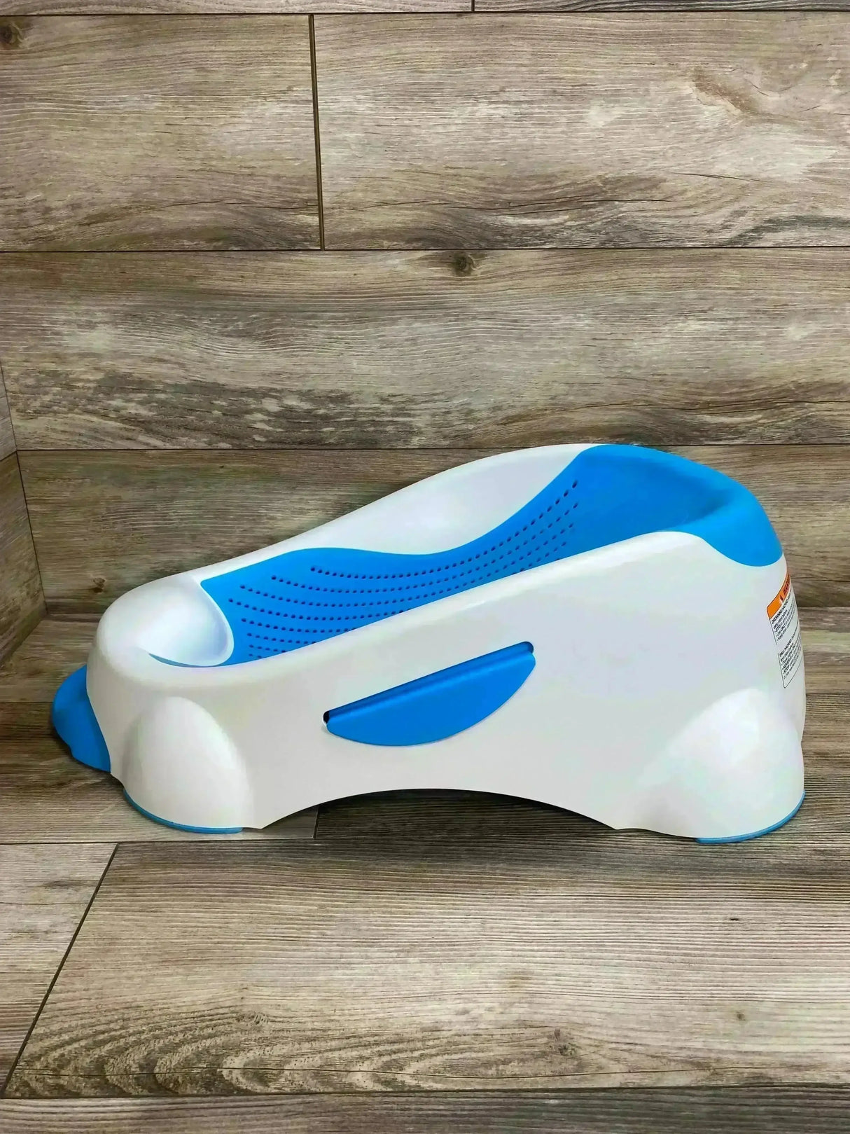 Side view of Munchkin Clean Cradle baby bathtub showcasing contoured blue padded insert, angled cradle shape, and non-slip feet on wood floor background