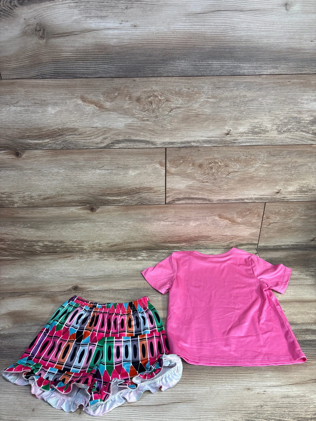 Flat-lay of toddler 2-piece set: pink tee and multicolor crayon-print ruffle shorts on light wood background, size 5T