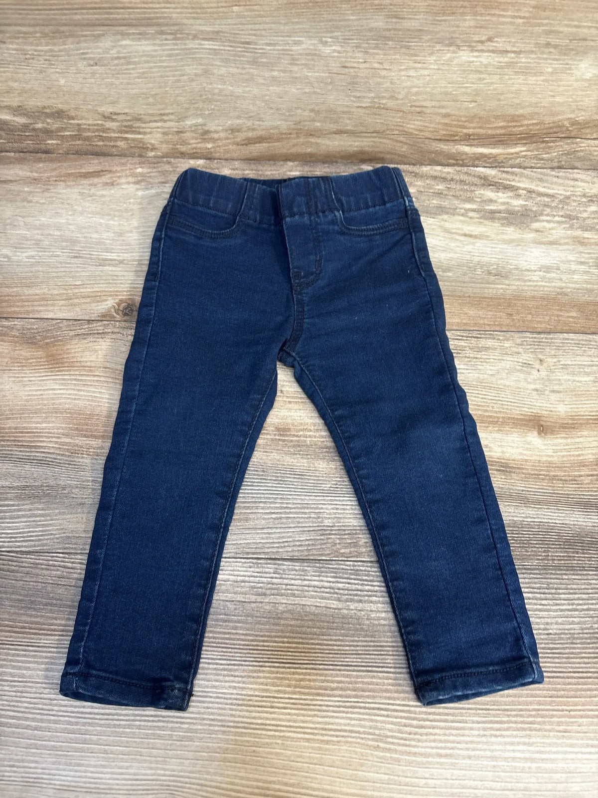 Flat lay of OshKosh navy pull on denim jeggings, size 18M, showing elastic waistband and tapered legs on wood floor