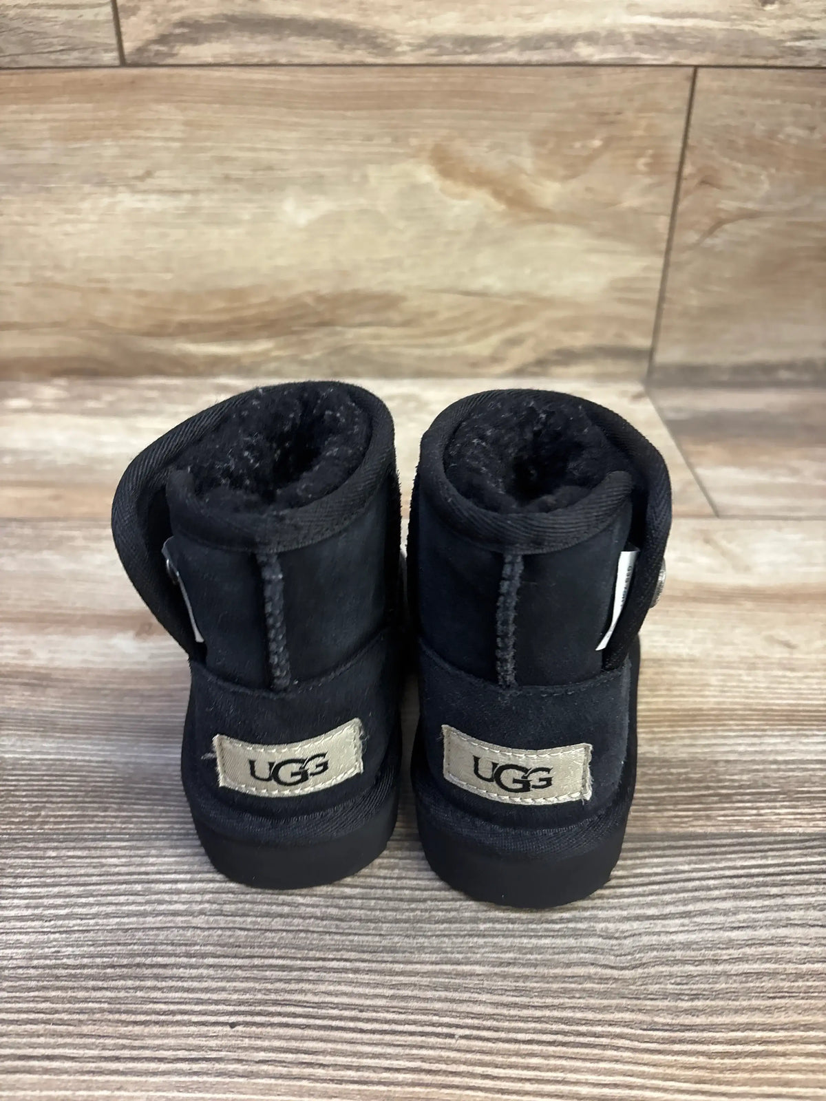 Rear view of black UGG Jona boots showcasing fleece-lined openings and UGG logo patches on a wood-look floor