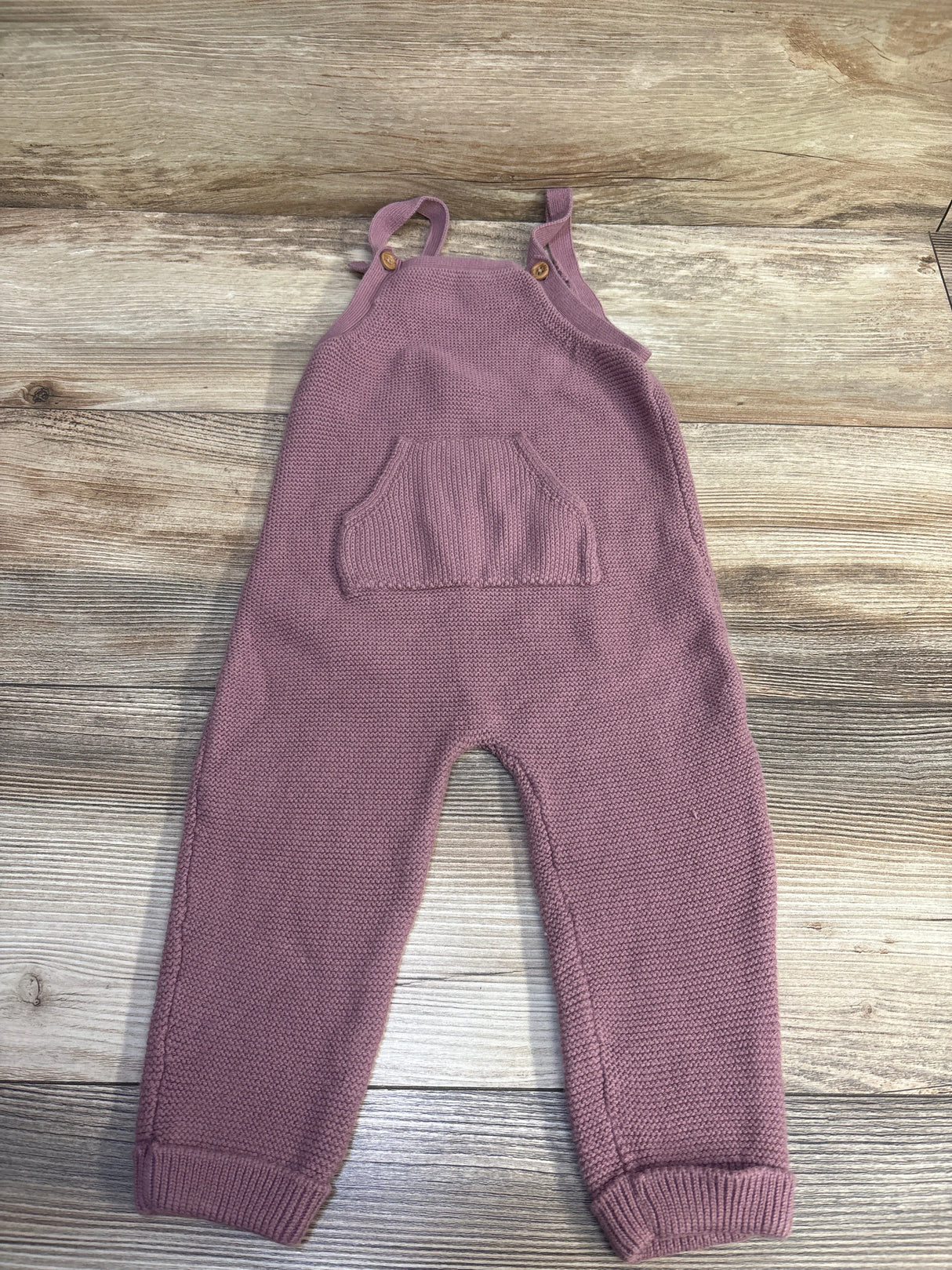 Front view of mauve knit overalls with buttoned straps, a front pocket, and ribbed cuffs on a wood background